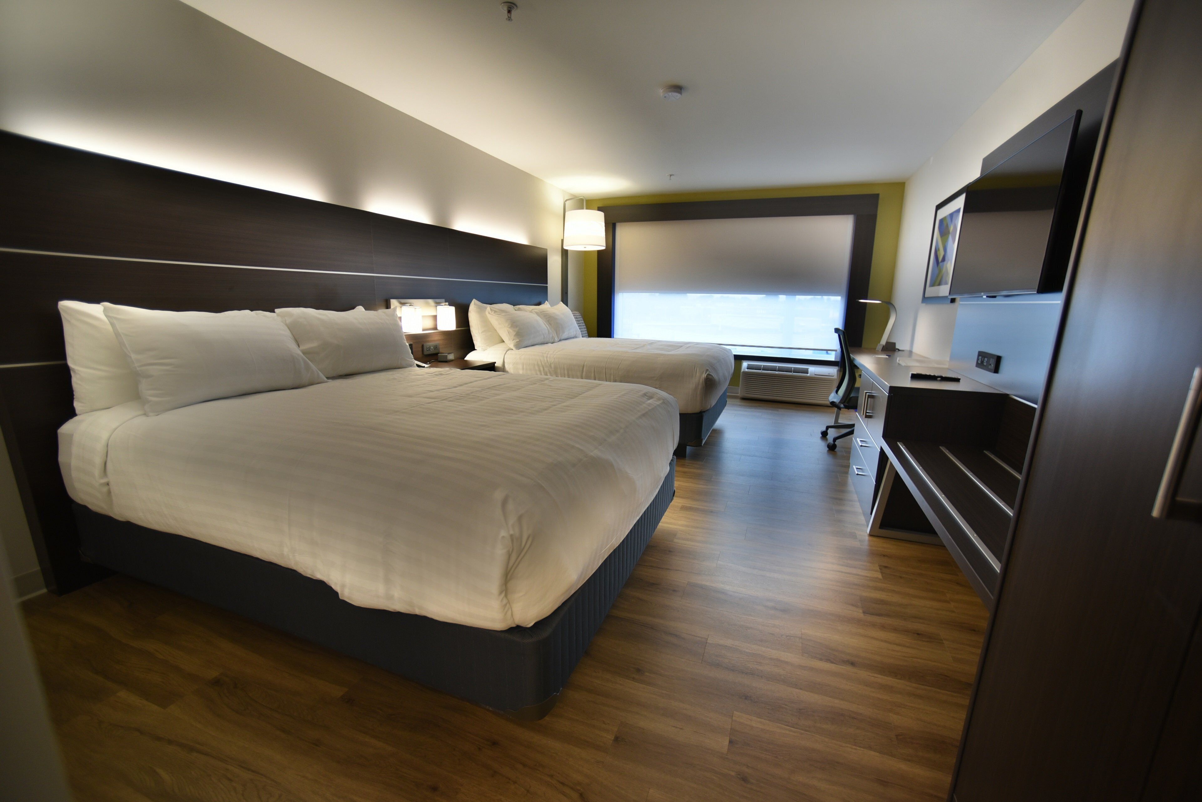 A modern hotel room with a large bed, a desk, a chair, a TV, and a window with blinds.