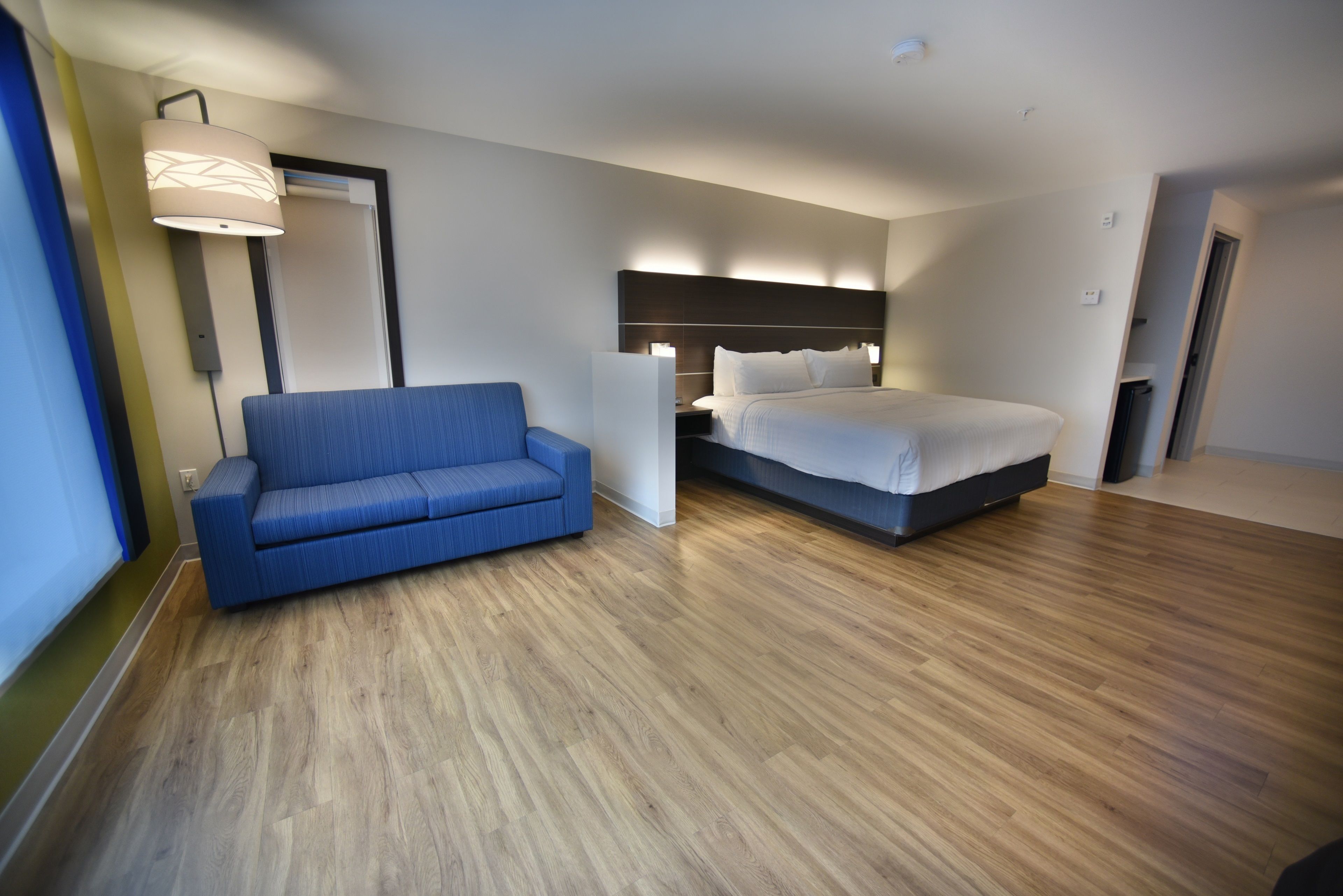 A modern hotel room with a blue sofa, a bed with a dark headboard, and a wooden floor.