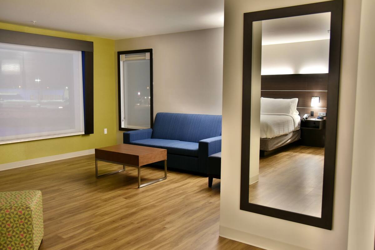 A hotel room with a large window, a blue sofa, a wooden coffee table, and a bed with a nightstand.