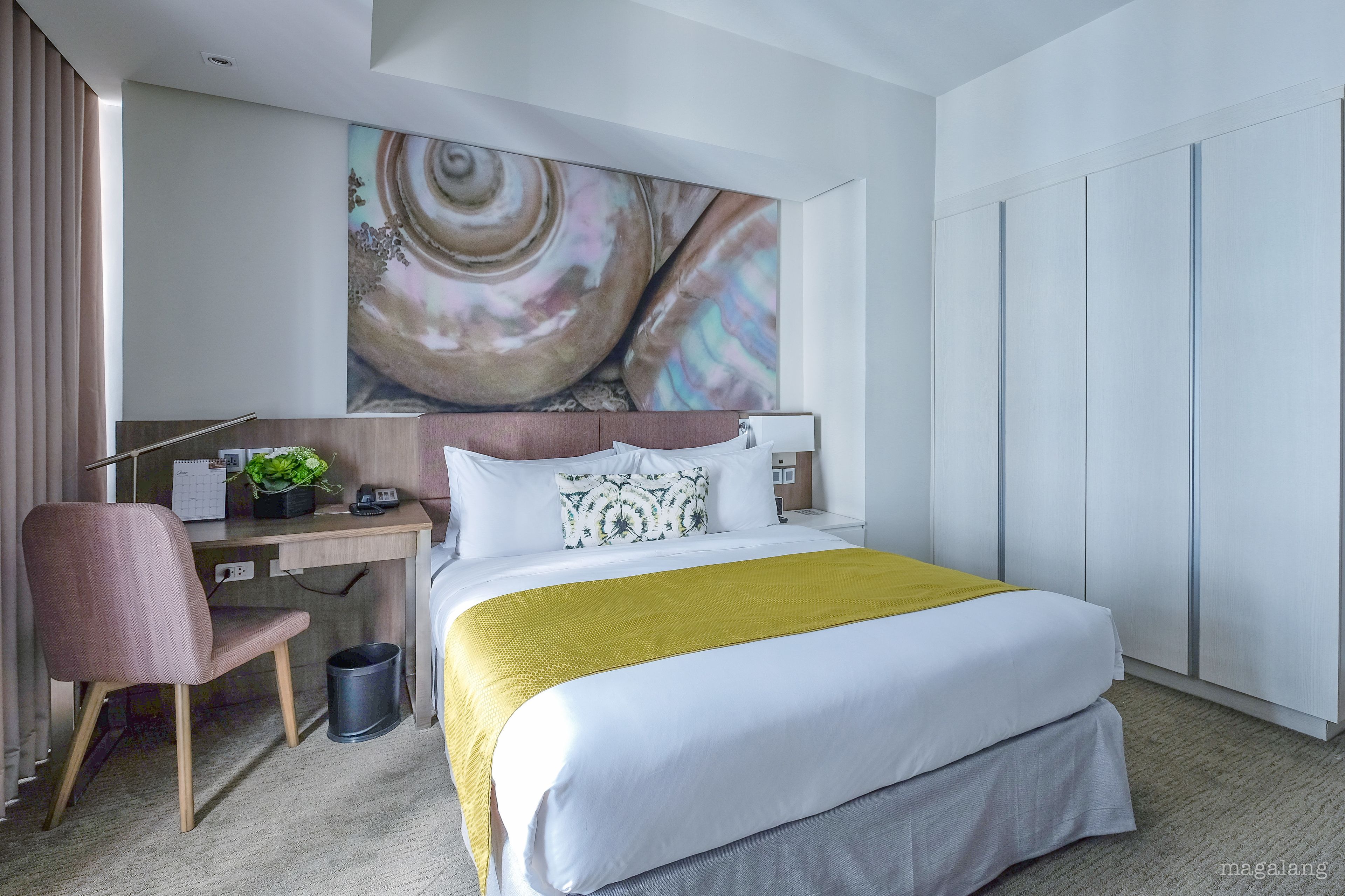 A hotel room with a bed, desk, chair, and a large abstract painting on the wall.