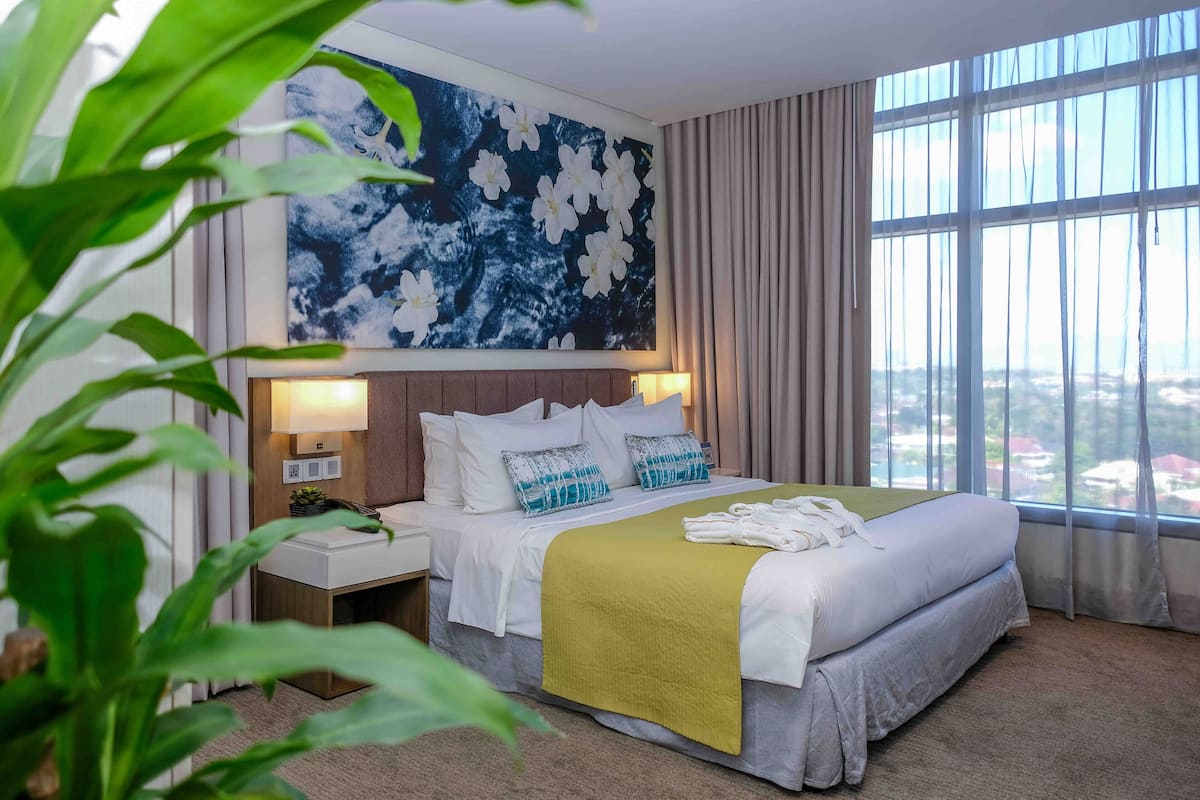 A hotel room with a bed, a nightstand, a lamp, a large window with curtains, and a painting on the wall.