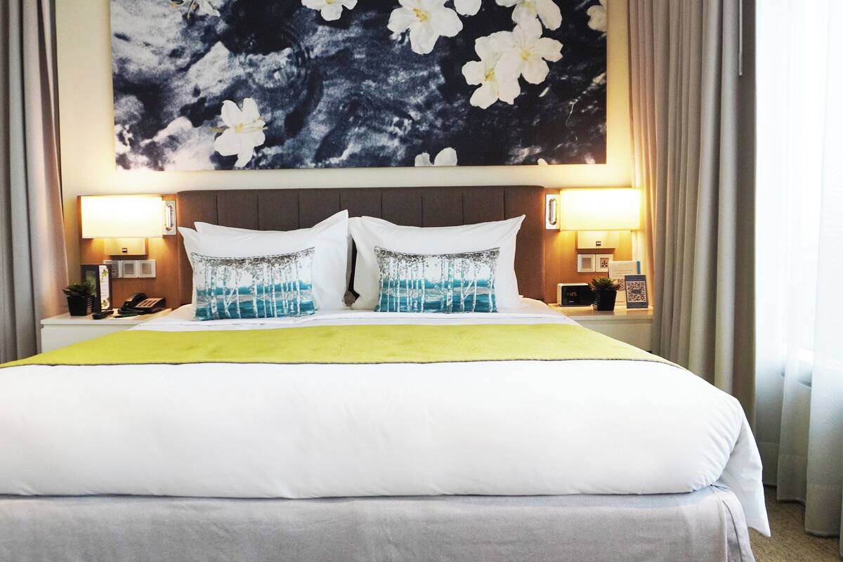A hotel room with a large bed, two bedside lamps, a nightstand with a phone and a plant, and a wall with a floral painting.