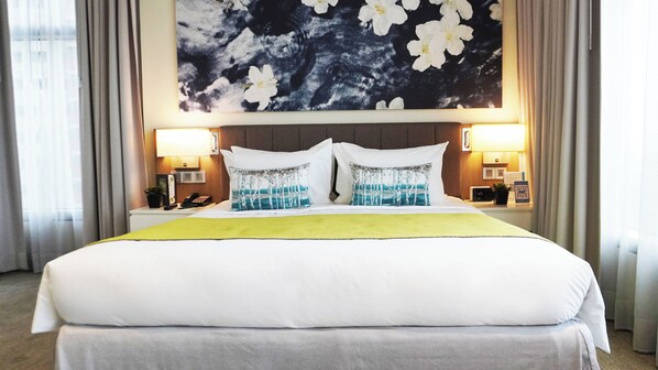A hotel room with a large bed, two bedside lamps, a nightstand with a phone and a plant, and a wall with a floral painting.