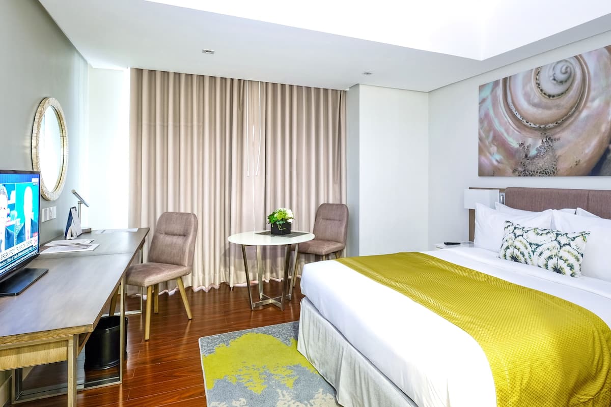 A hotel room with a bed, a desk, a chair, a small table with a plant, and a large abstract painting on the wall.