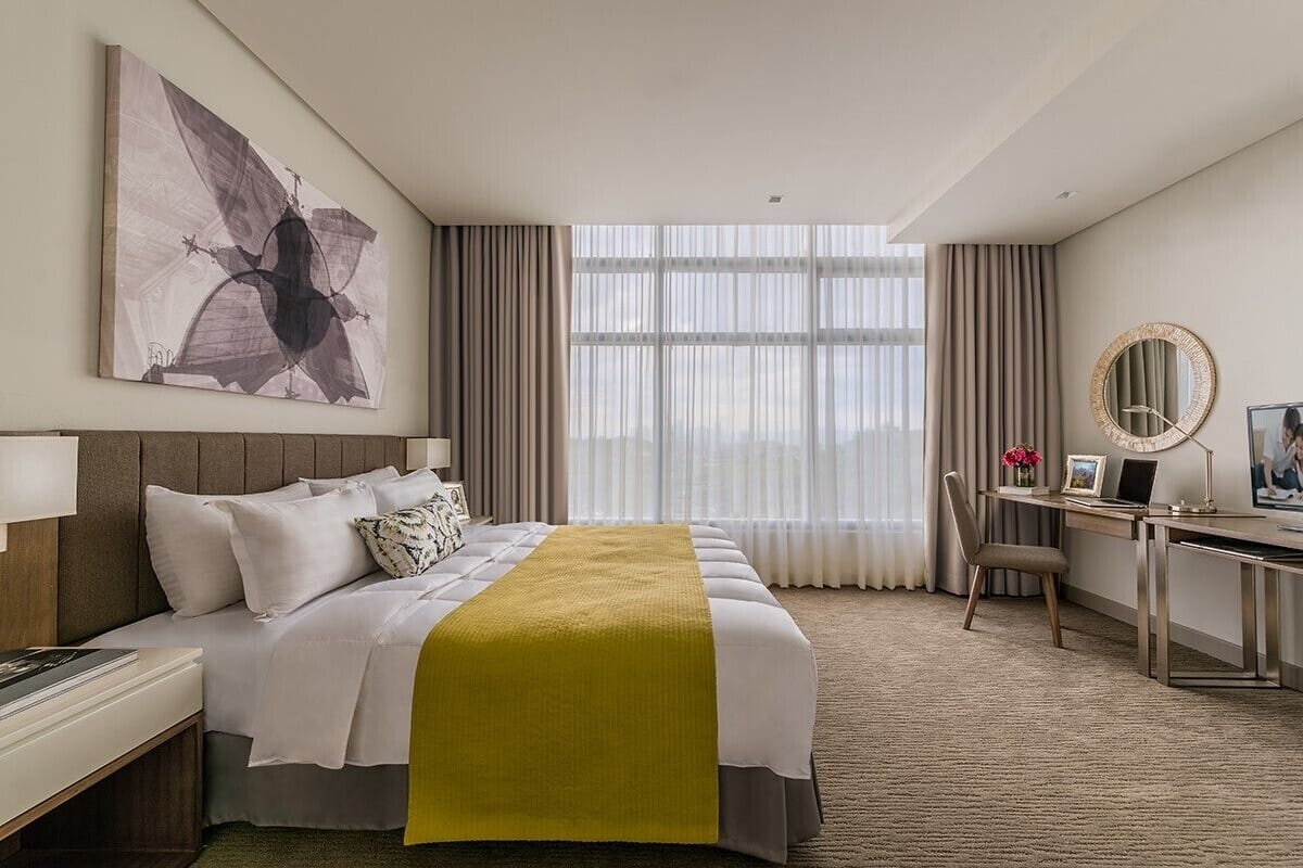 A hotel room with a large bed, a desk with a chair, a large window with curtains, and a painting on the wall.