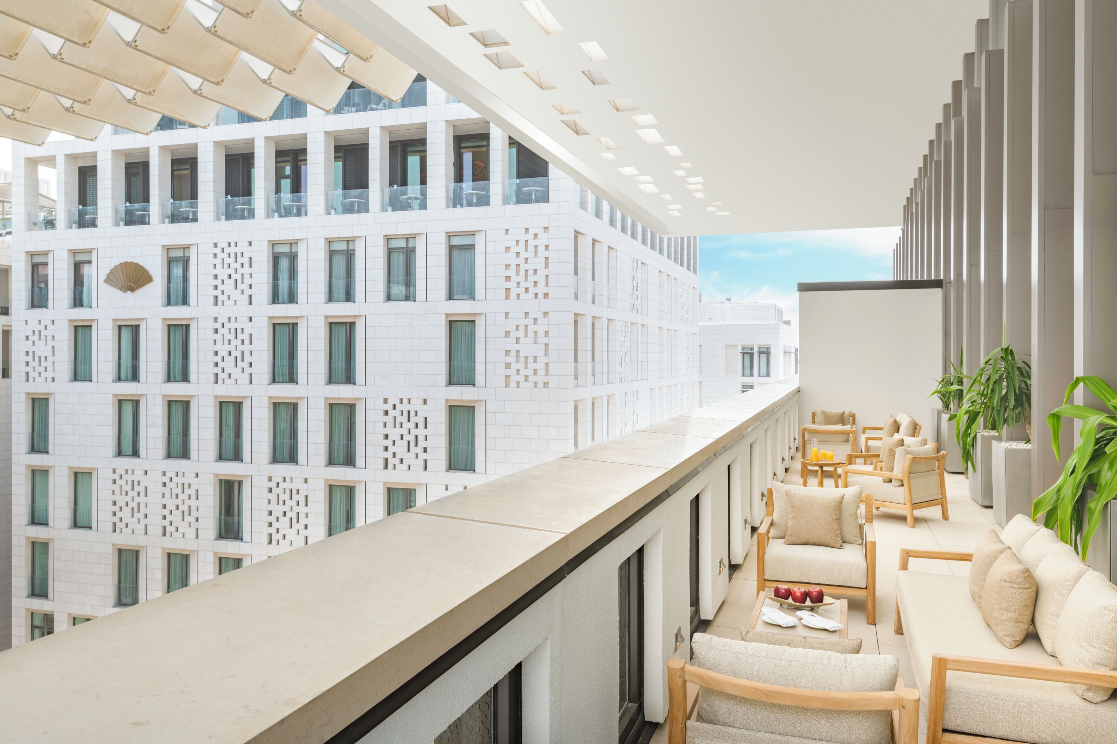 Een modern balkon van een hotel met uitzicht op een wit gebouw, bemetoleerd zitmeubilair en potplanten.