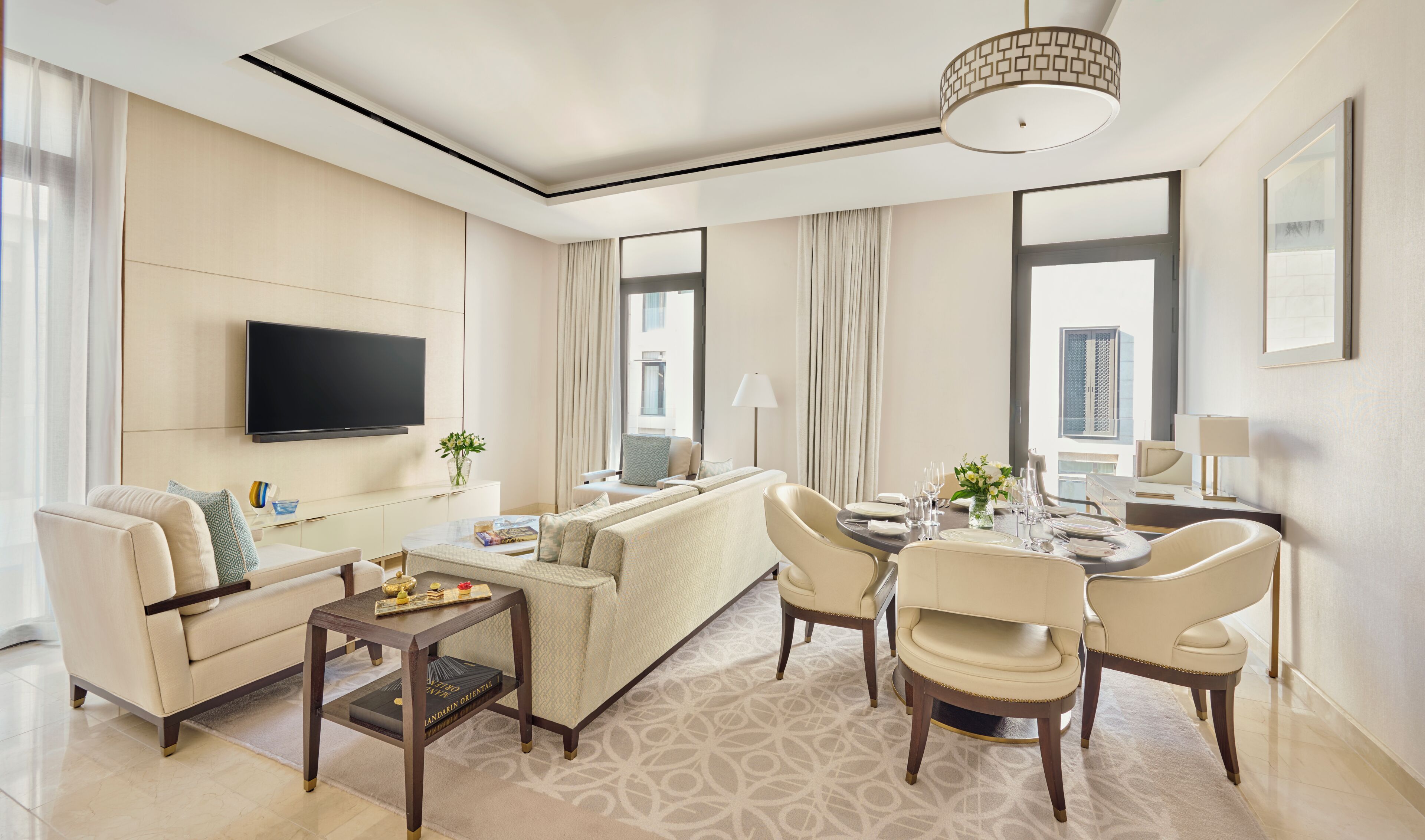 Habitación de hotel moderna con sofá, una mesa de comedor para dos personas, televisor montado en la pared y ventanas grandes con cortinas.