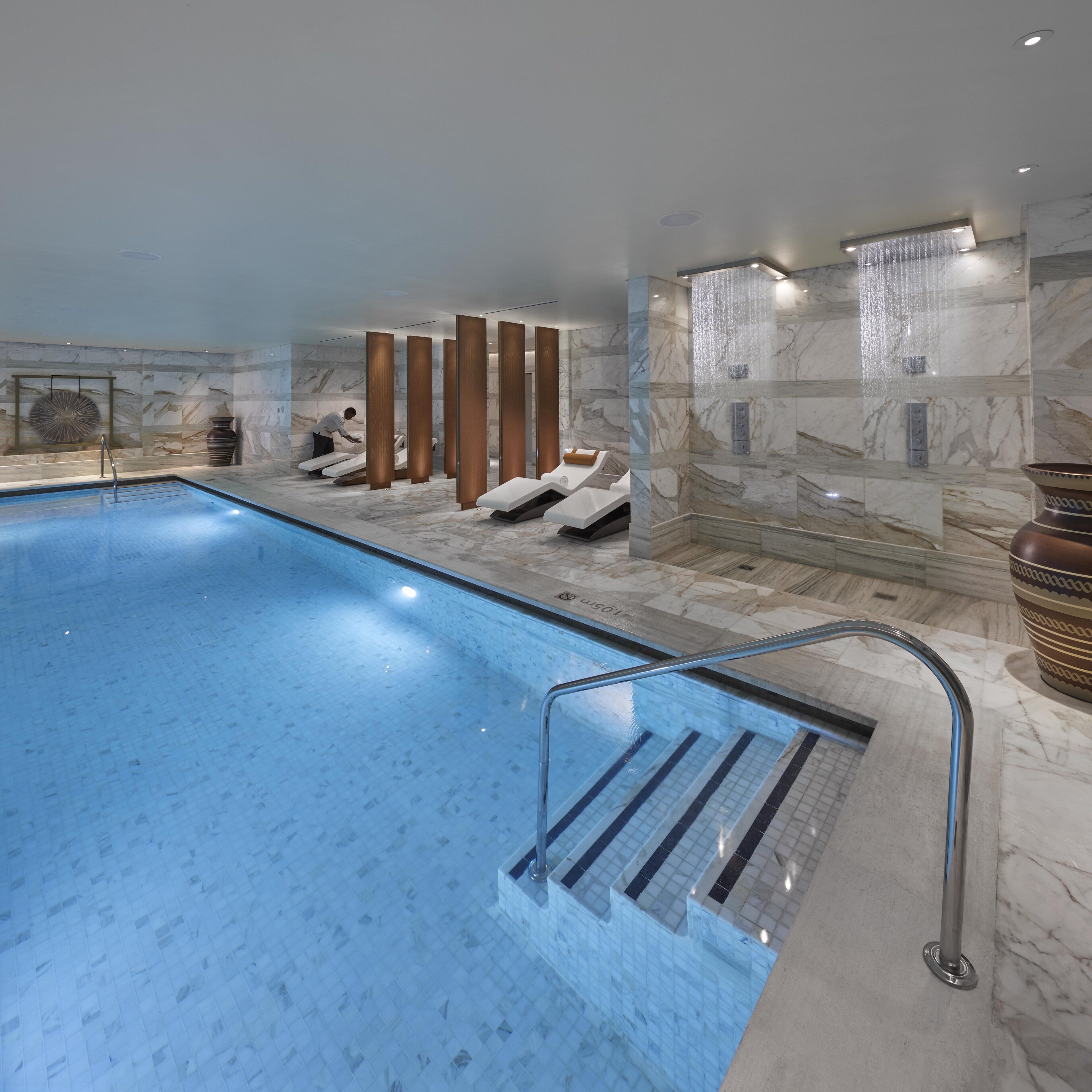 A modern indoor swimming pool with a clear blue water surface, surrounded by marble walls and a seating area with loungers.