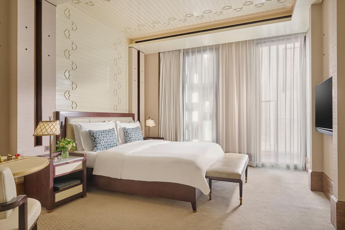 A hotel room with a large bed, bedside tables, a TV, and a view of the outdoors through sheer curtains.