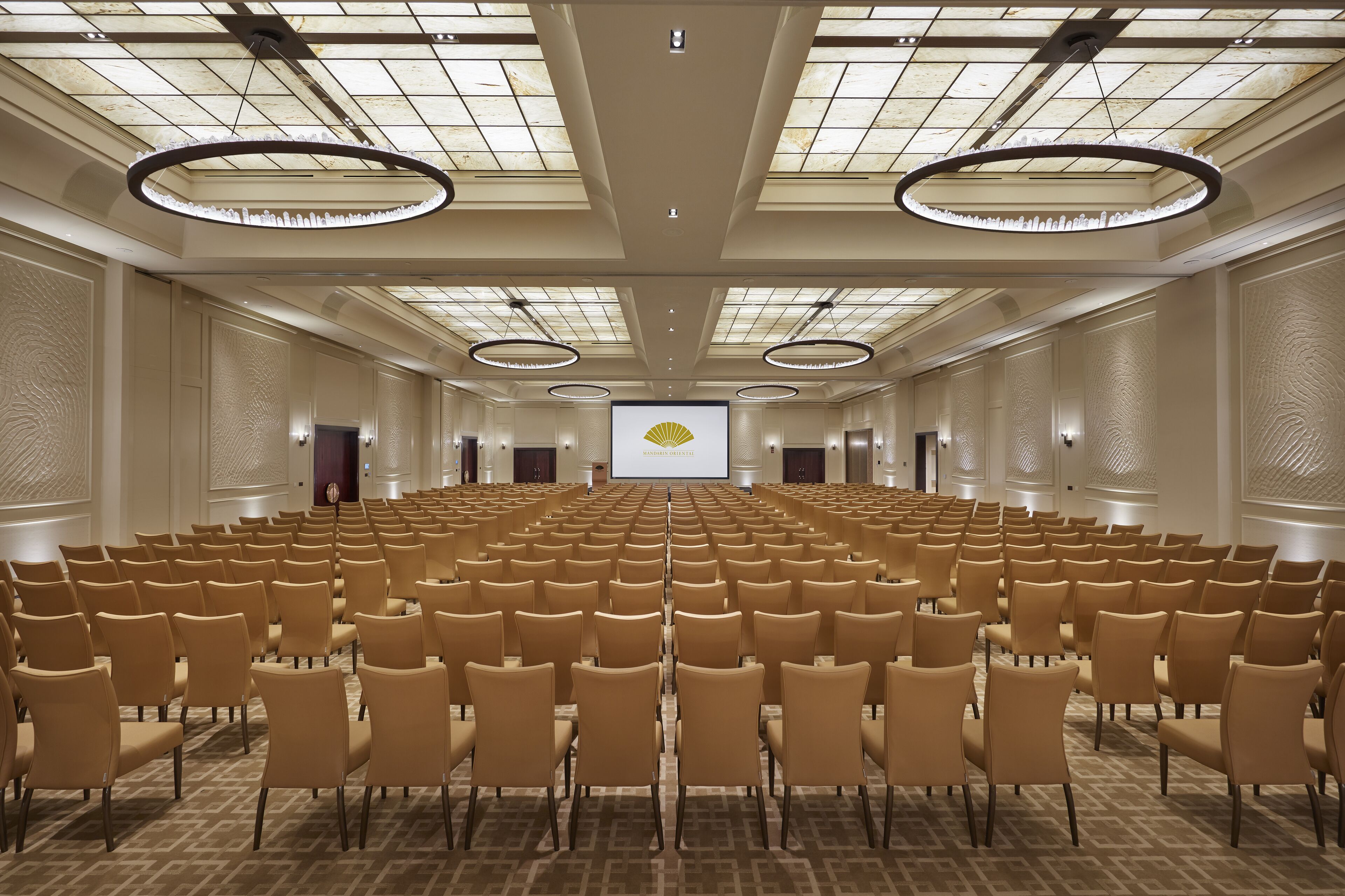 Un salón de conferencias con filas de sillas, una pantalla de proyector y un techo con tragaluces y luminarias circulares.