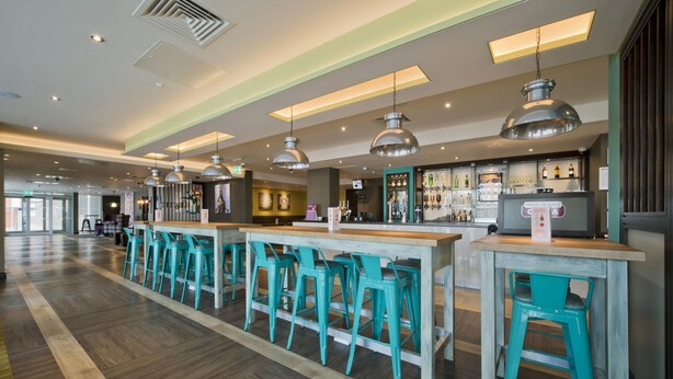 A bar area with wooden tables and teal chairs, a bar counter, and pendant lighting. - Premier Inn Leeds Cc - Whitehall Road (Leeds)