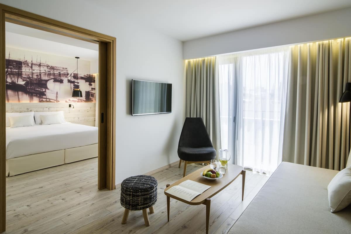 Quarto de hotel com cama, televisão, mesa de jantar com uma tigela de frutas, cadeira e janela com cortinas.