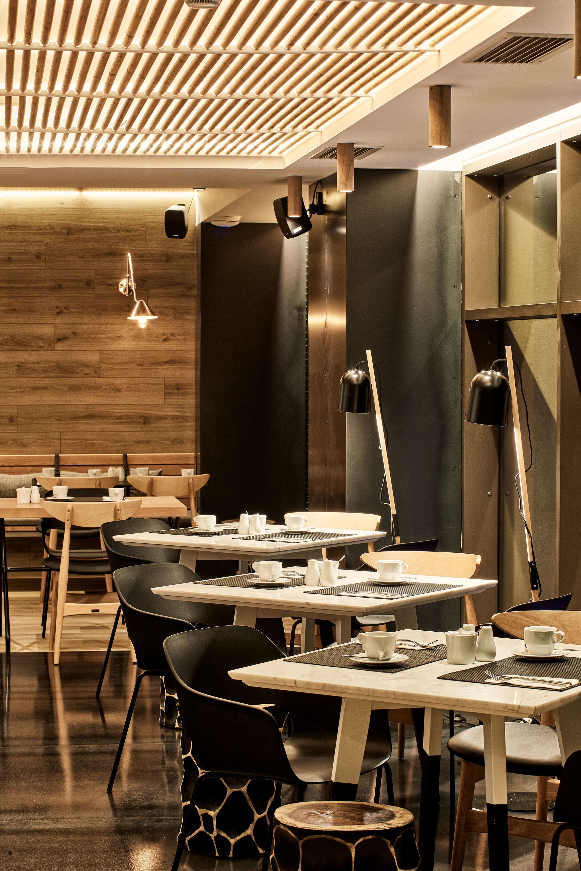 Interior moderno de restaurante con paneles de techo de madera, sillas negras y de madera, y mesas dispuestas con vajilla blanca.
