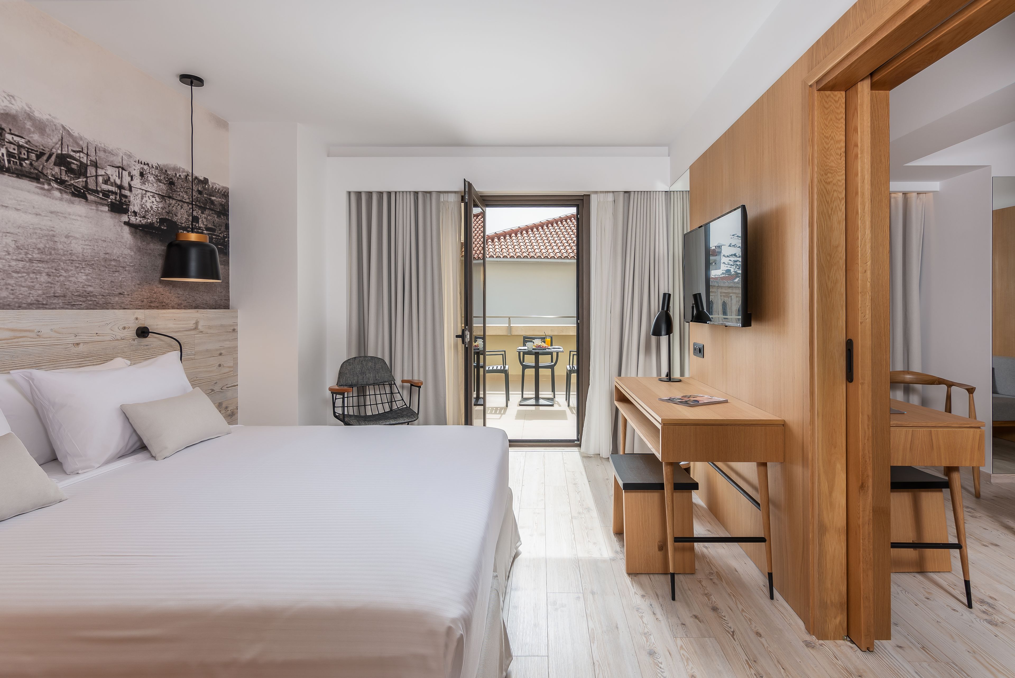 A hotel room with a bed, a desk, a chair, a TV, and a balcony with curtains.