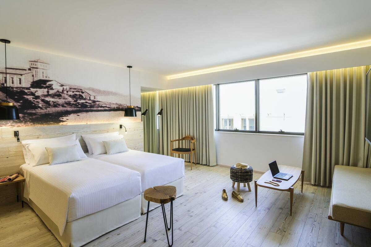A hotel room with two beds, a desk, a chair, a small stool, and a large window with curtains.