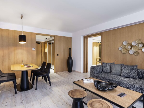 A modern living room with a wooden floor, a round dining table with chairs, a sofa, and a coffee table with a remote control.