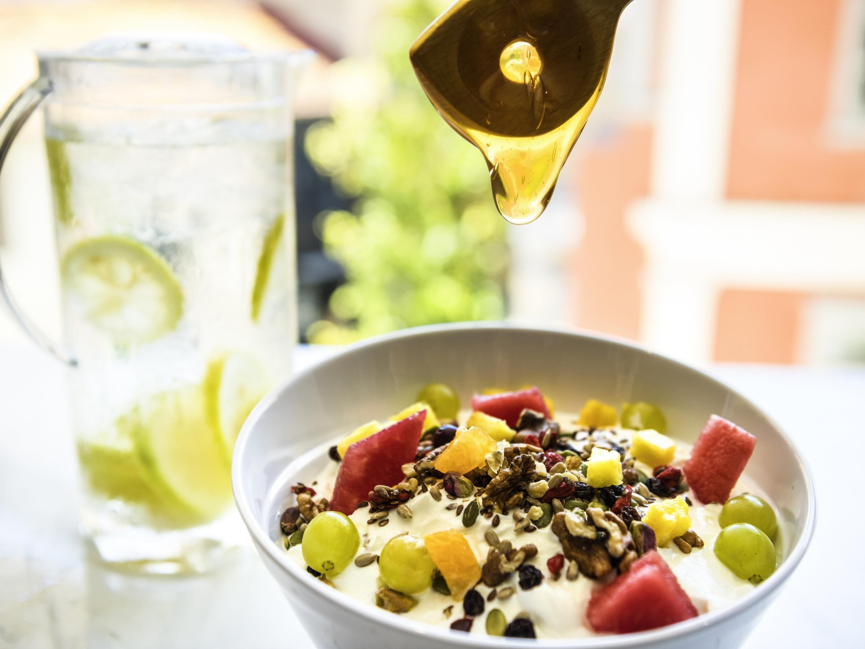 A bowl of yogurt topped with fruits and nuts, a glass of iced drink with lime, and a pitcher in the background.