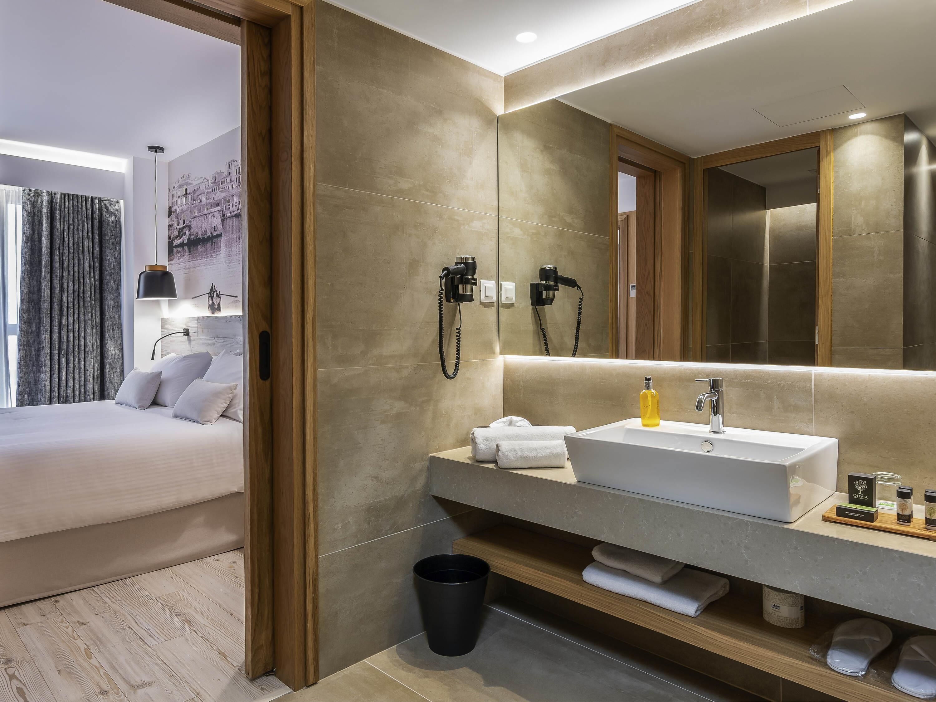 A modern hotel room with a large bed, a vanity with a sink, and a mirror.