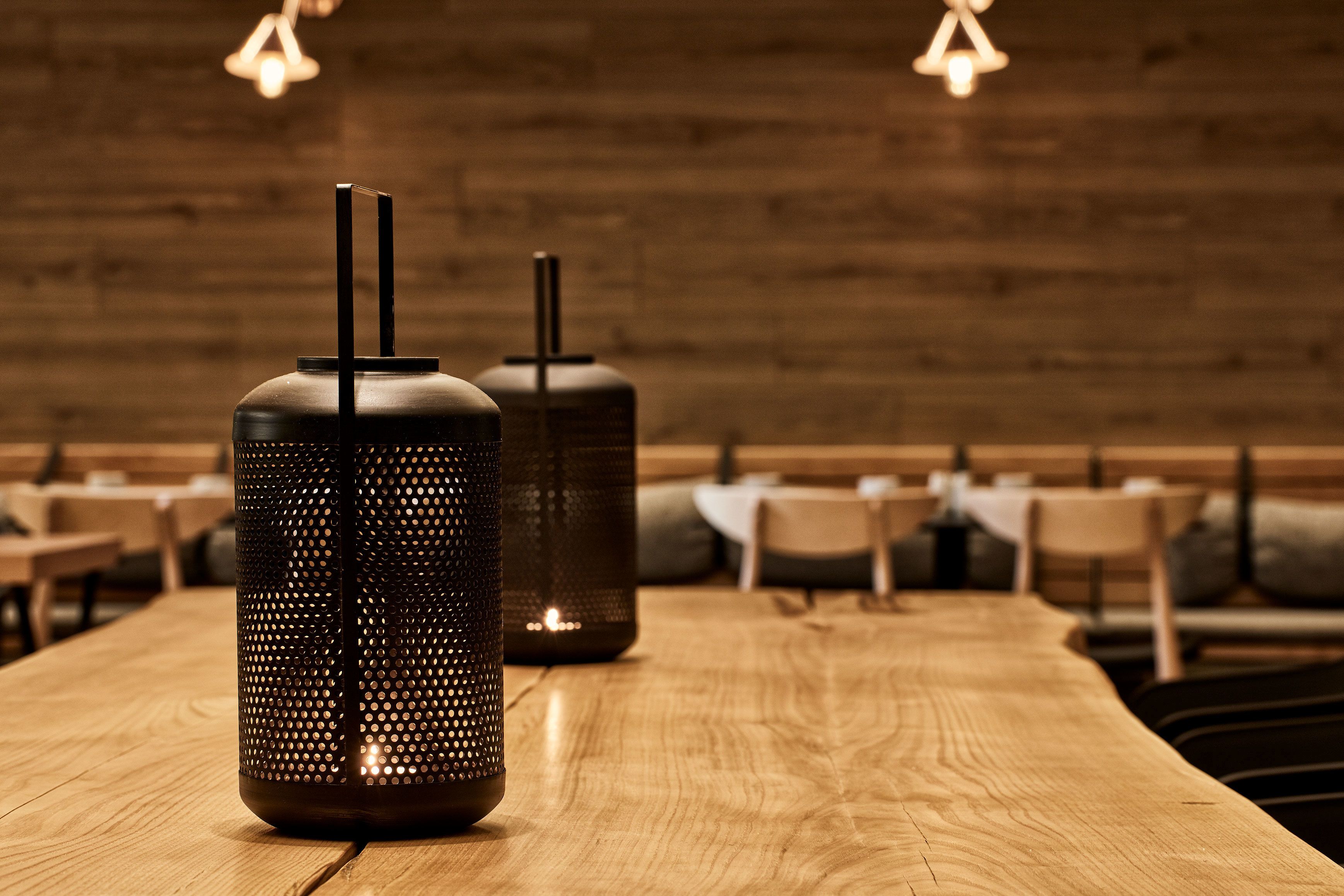 Two tall, dark-colored, perforated candle holders on a wooden table in a restaurant setting with wooden walls and hanging lights.
