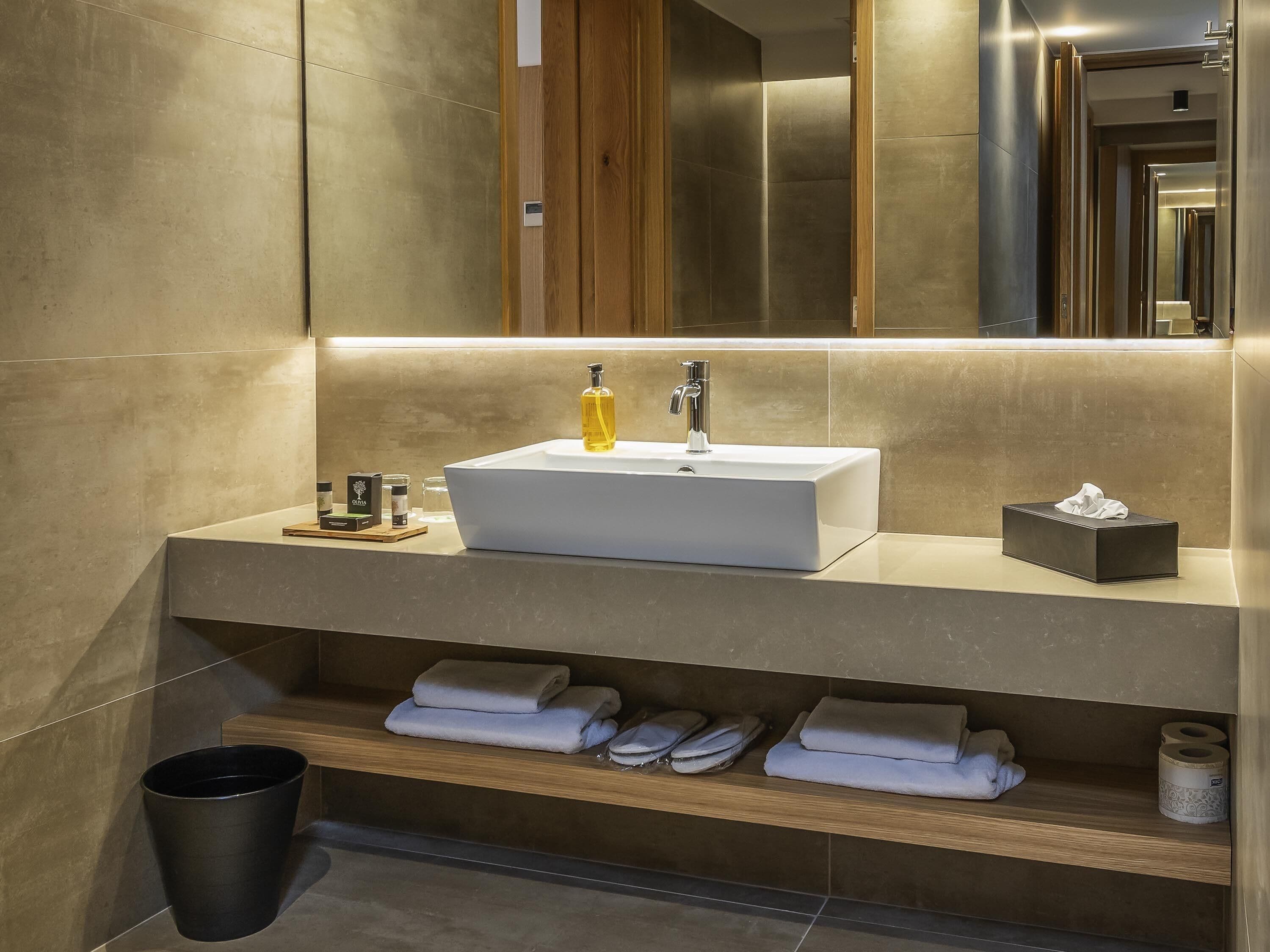 A modern bathroom with a white sink, a mirror, and a shelf with towels and toiletries.