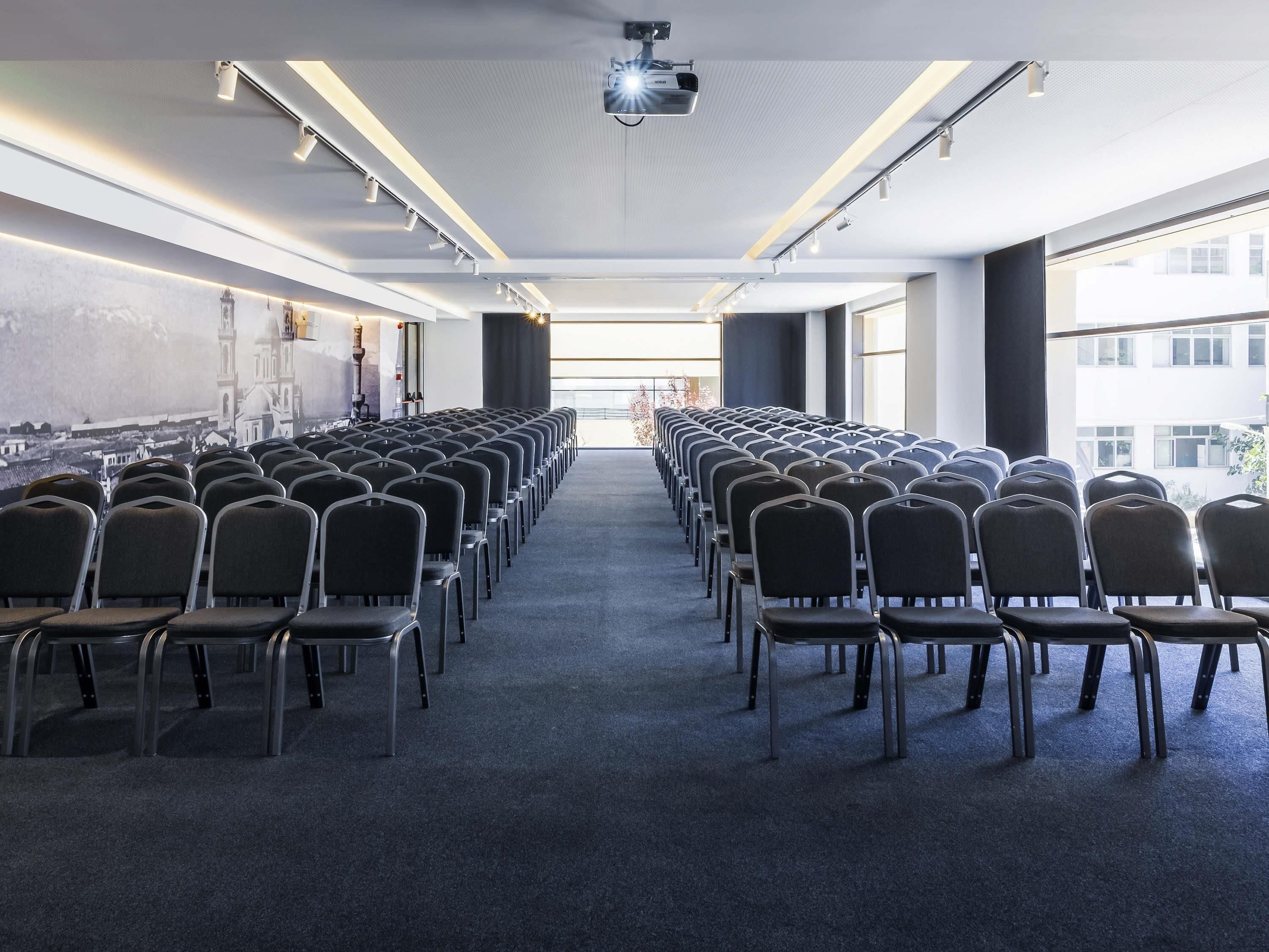 Un salón de conferencias con filas de sillas, un proyector y ventanas amplias.