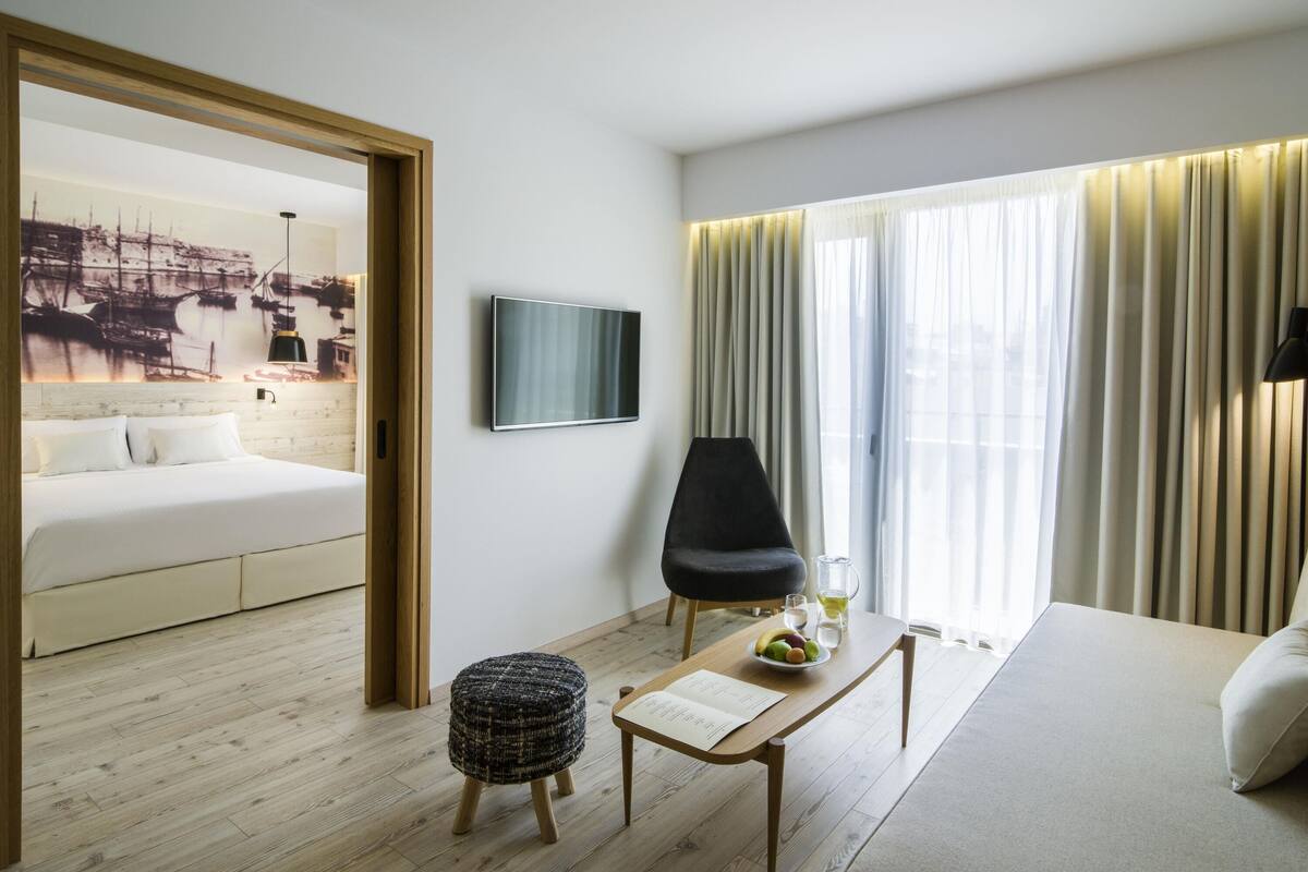 A hotel room with a bed, a TV, a dining table with a bowl of fruit, a chair, and a window with curtains.