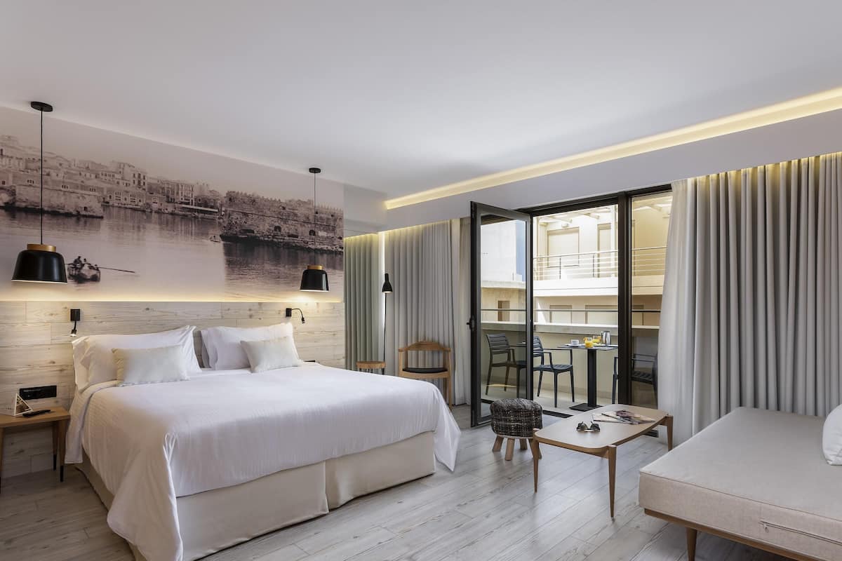 Quarto de hotel moderno com uma cama grande, uma mesa pequena, um sofá e uma varanda com cortinas.