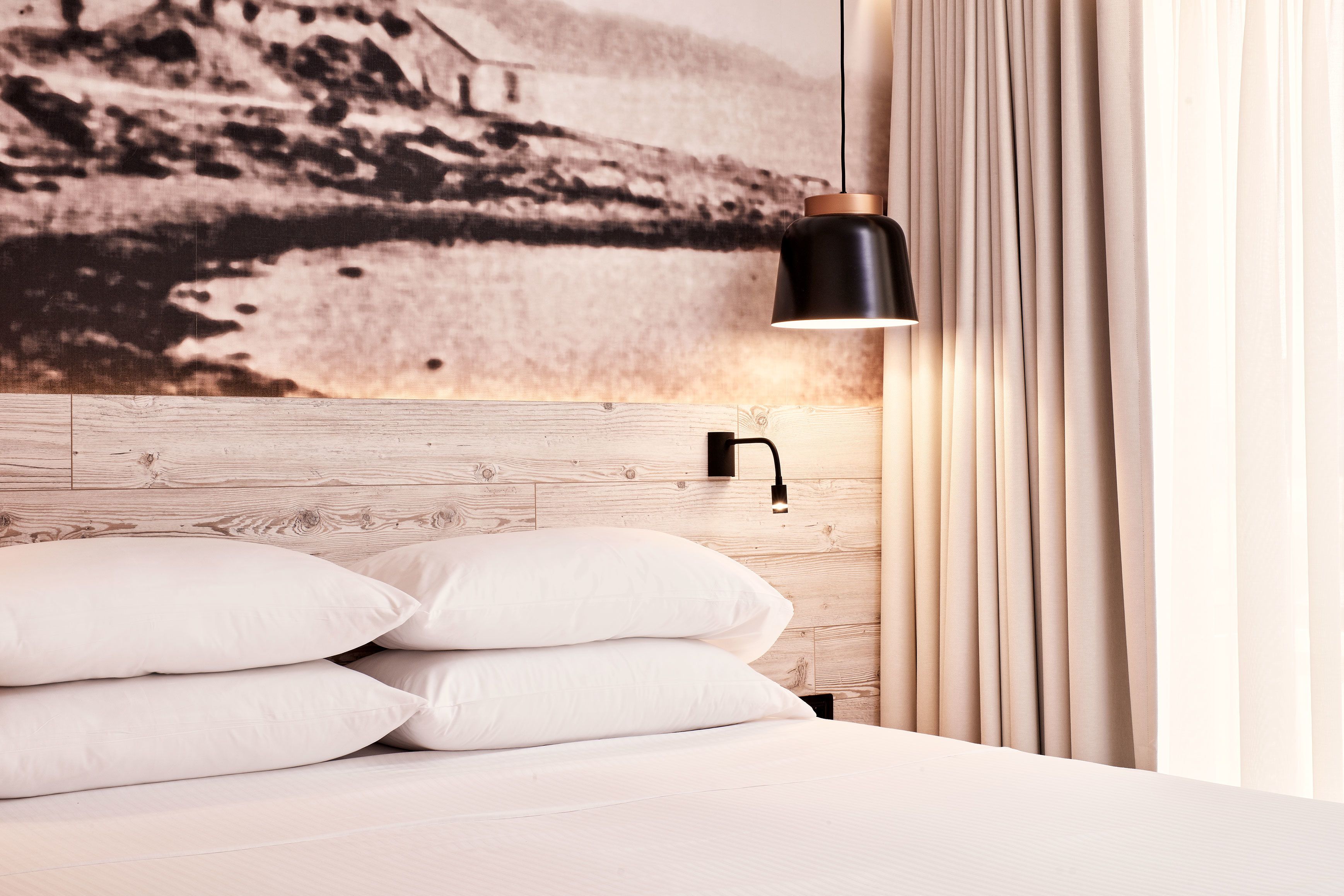 Una cama bien hecha con ropa blanca y almohadas, una lámpara negra de pared y un cabecero de madera.