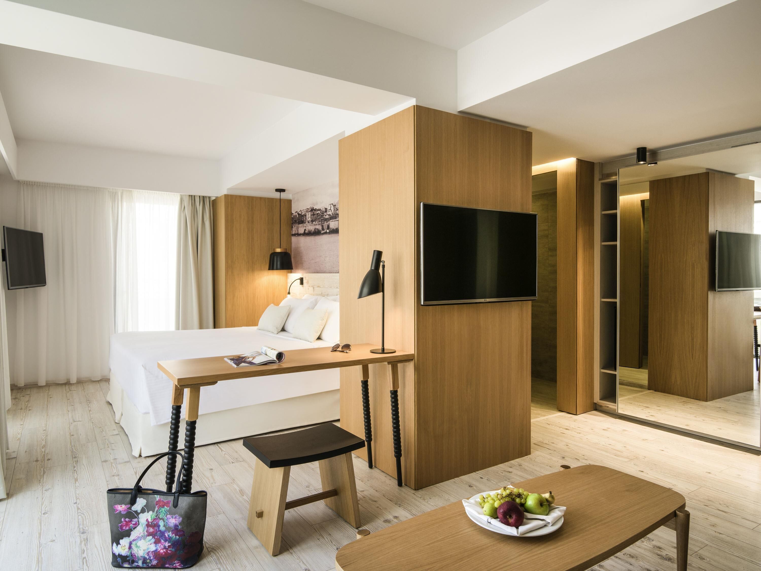 A hotel room with a bed, desk, chair, TV, and a dining table with fruit.