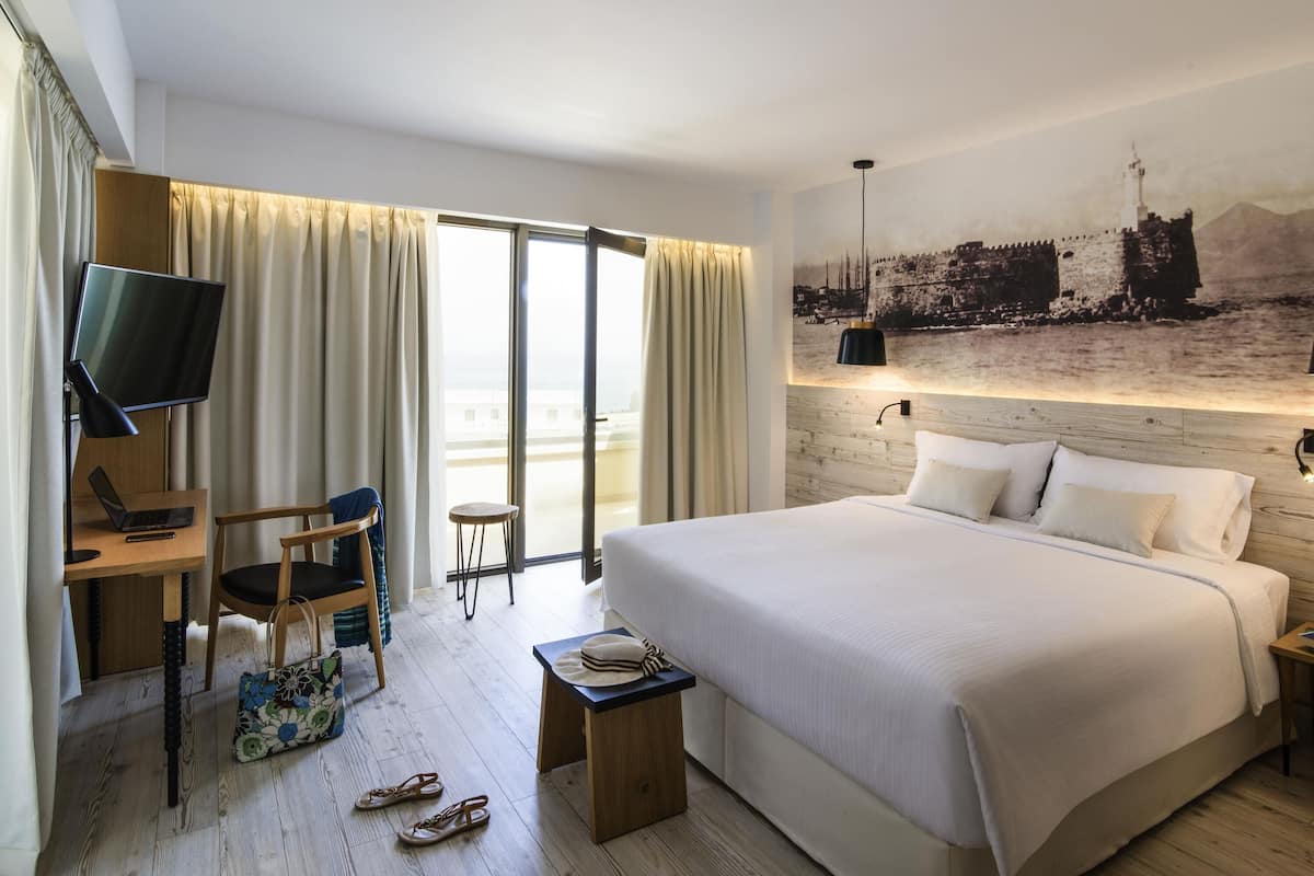 Quarto de hotel com uma cama grande, uma escrivaninha com um computador, uma cadeira, uma mesinha com um chapéu e uma televisão.