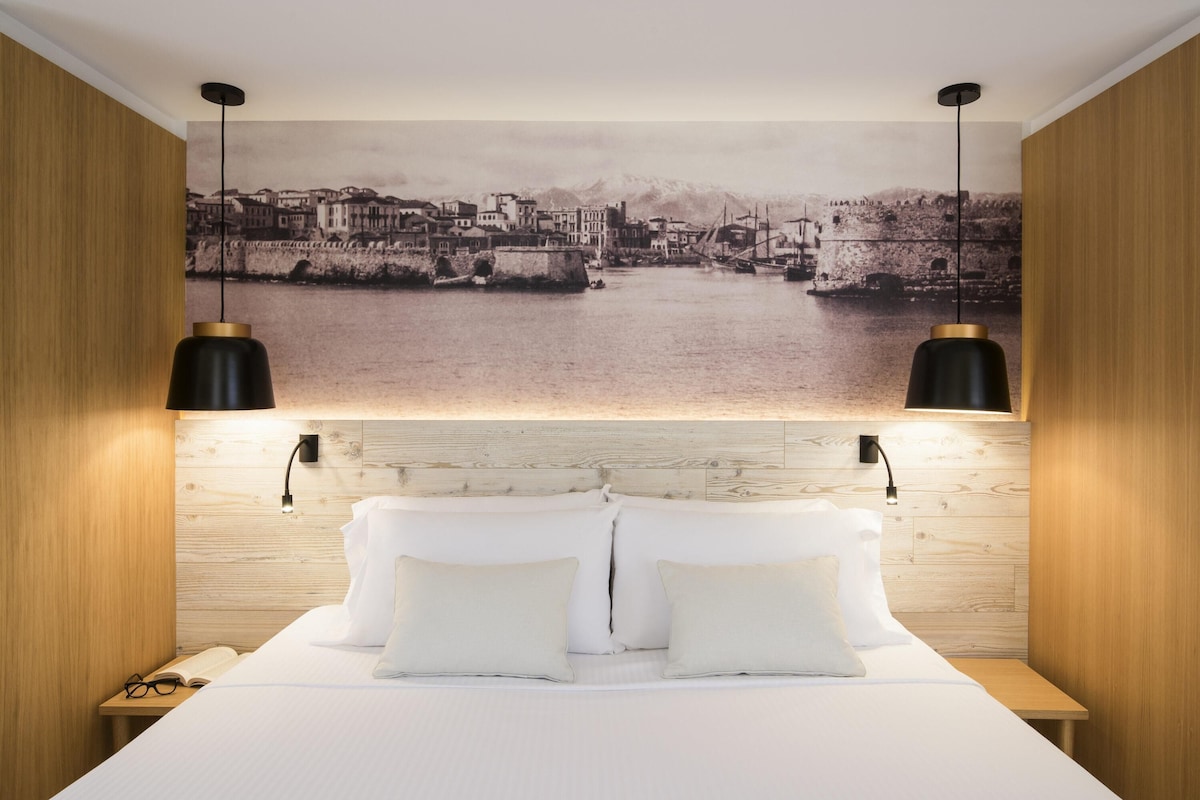 A bedroom with a large bed, two bedside lamps, and a cityscape mural on the wall.