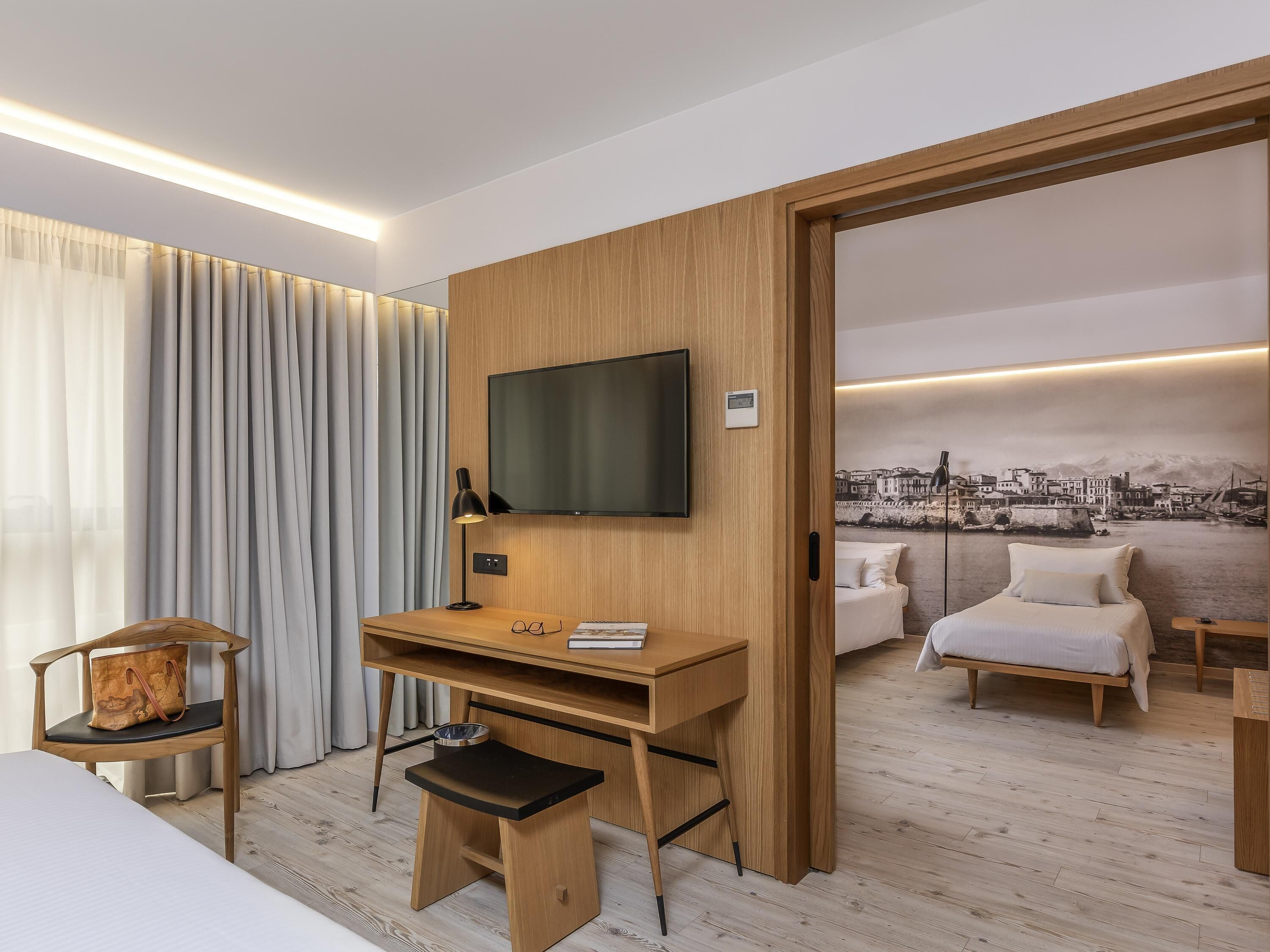 Habitación de hotel con un escritorio de madera, televisor, cama, una silla y vistas a un paisaje urbano a través de un ventanal grande.