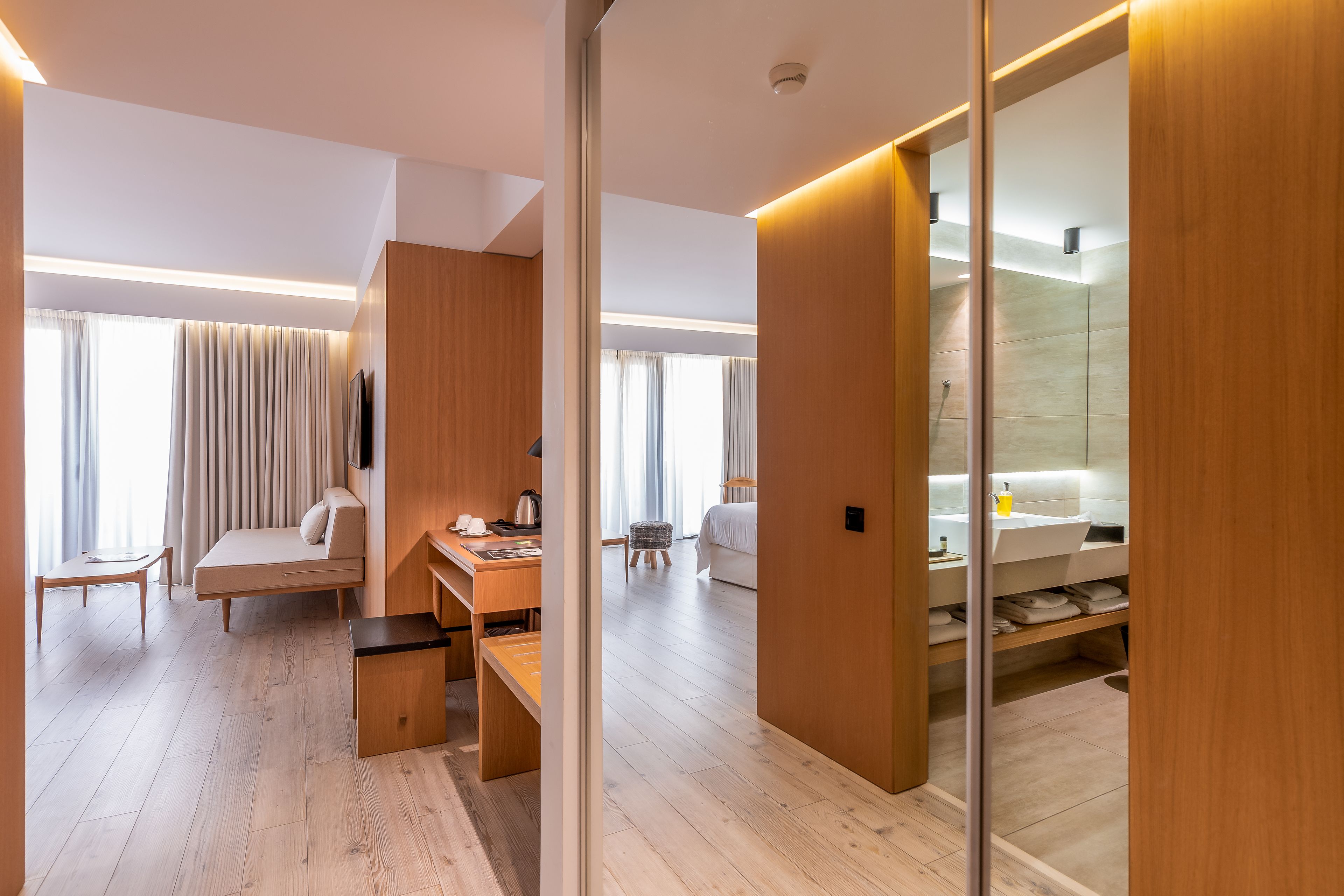 Une chambre d’hôtel moderne avec un plancher en bois, un canapé, un bureau, une chaise, une petite table, un miroir et une salle de bain équipée d’un lavabo et d’étagères.