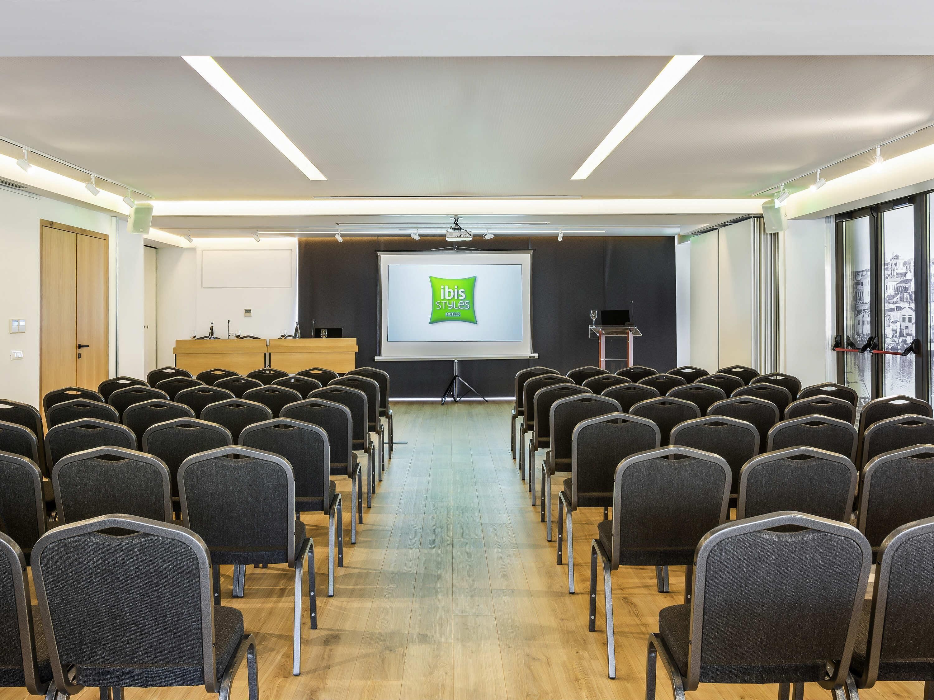 Een vergaderzaal met rijen stoelen, een spreekgestalte, een scherm met het logo van 'ibis Styles Hotels' en een stadszicht door grote ramen.