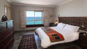 Un dormitorio con una cama grande, un aparador de madera, una mesita de noche, una lámpara, una ventana con contraventanas y vistas al océano.