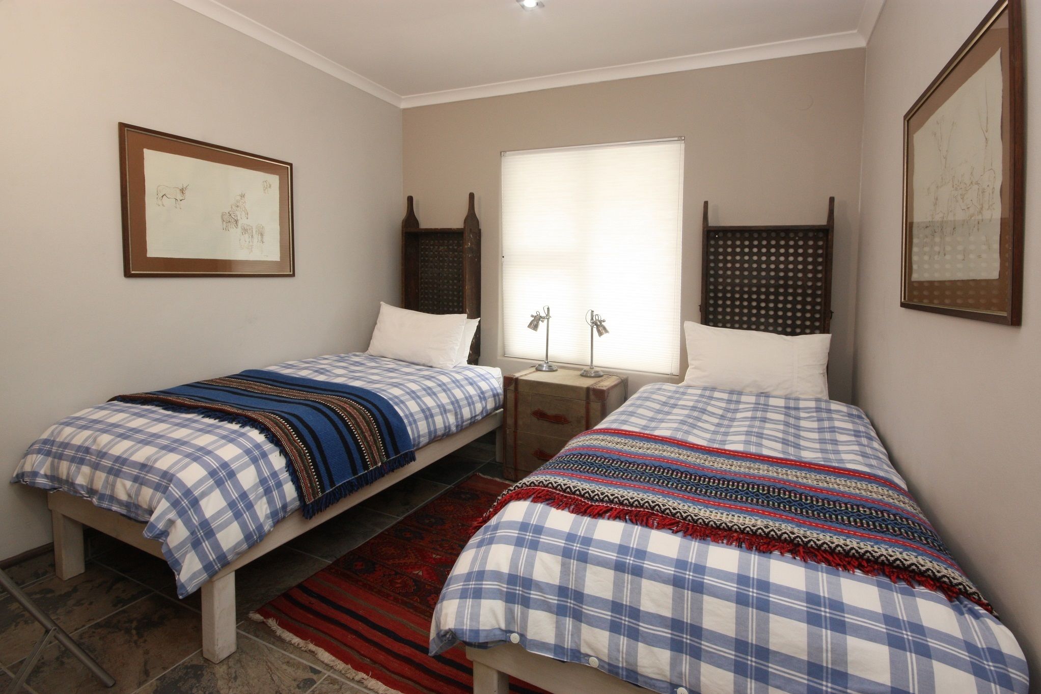 Een kamer met twee bedden, een raam met jaloezieën, een lijst met een afbeelding aan de muur en een nachtkastje met een lamp.