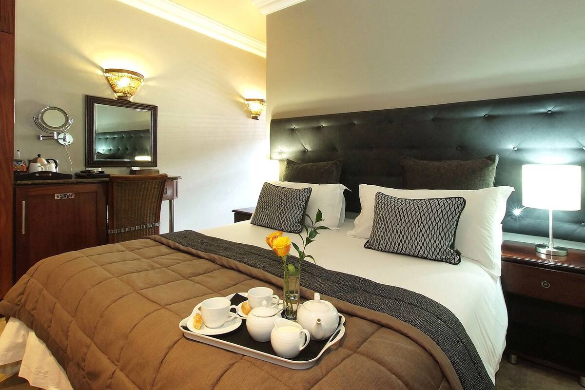 A hotel room with a large bed, a bedside table with a lamp, a dresser, a mirror, and a tray with breakfast items.