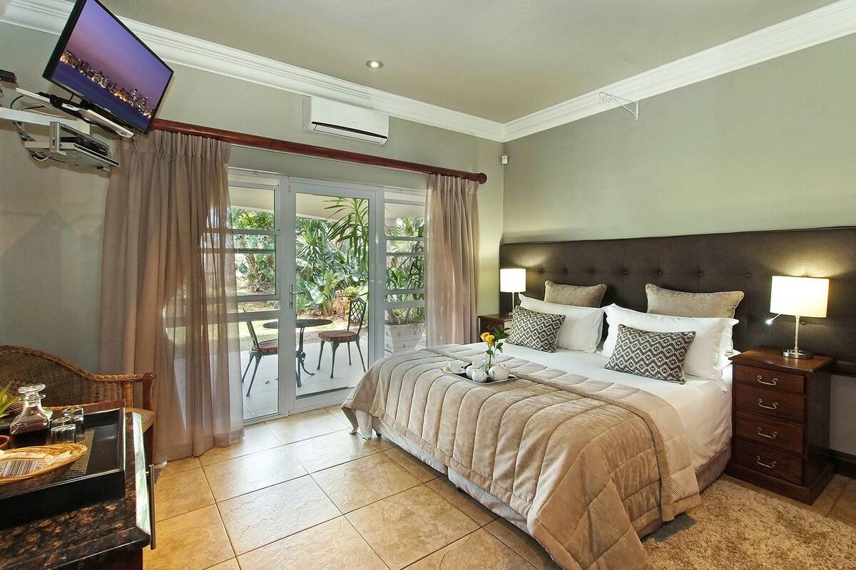 A hotel room with a bed, bedside lamps, a TV, a dining area with chairs, and a view of greenery through the glass doors.