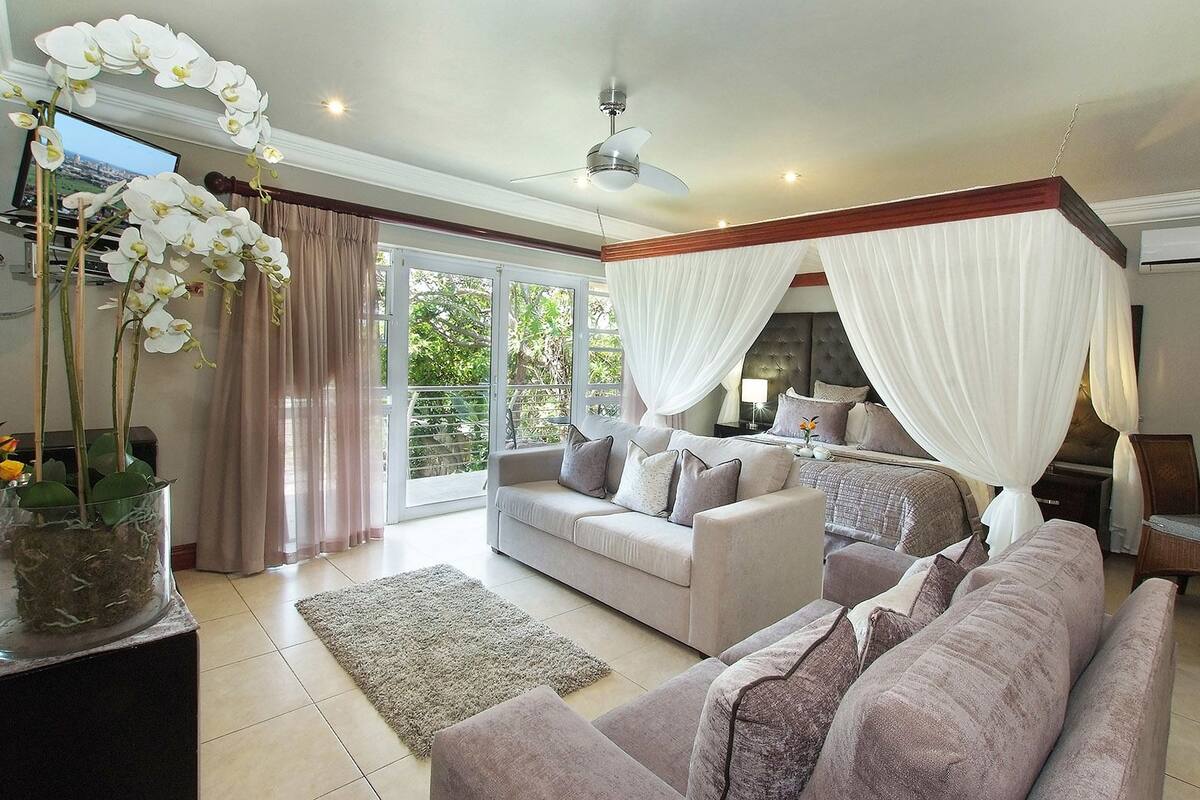 A spacious living room with a large sofa, a smaller sofa, and a coffee table. There is a large window with curtains, a ceiling fan, and a potted plant with white flowers.