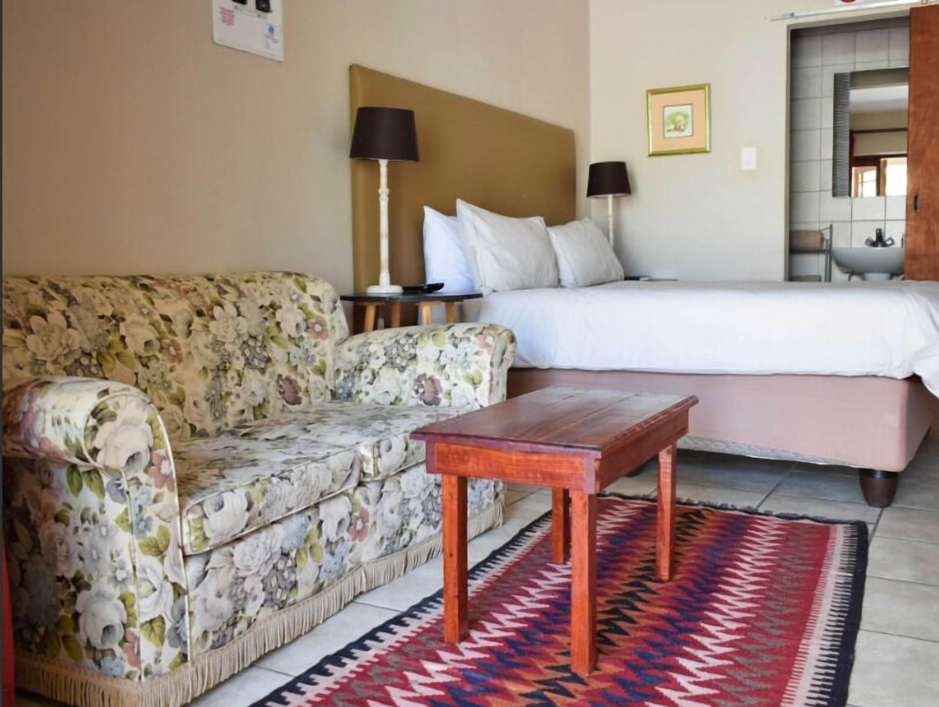 A hotel room with a floral sofa, a wooden table, a bed with white bedding, and a patterned rug.