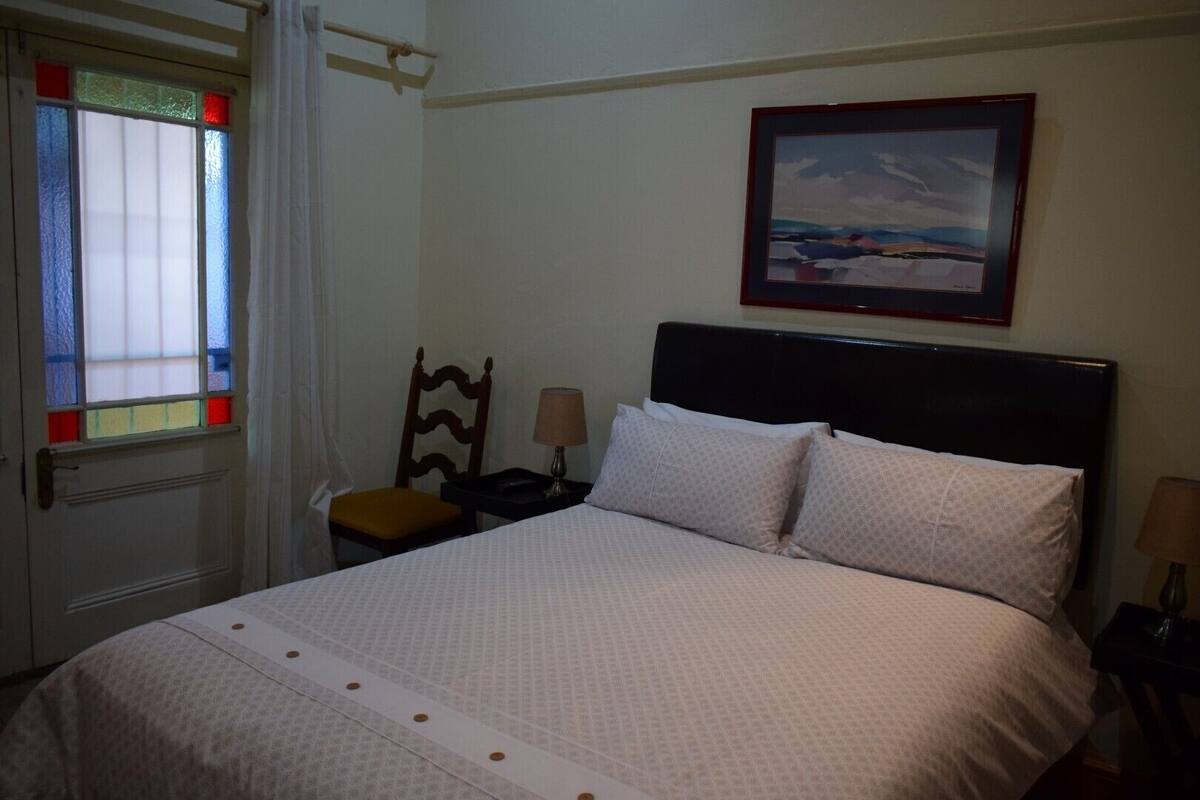 A neatly made bed with a headboard, a chair, a bedside table with a lamp, and a window with stained glass.