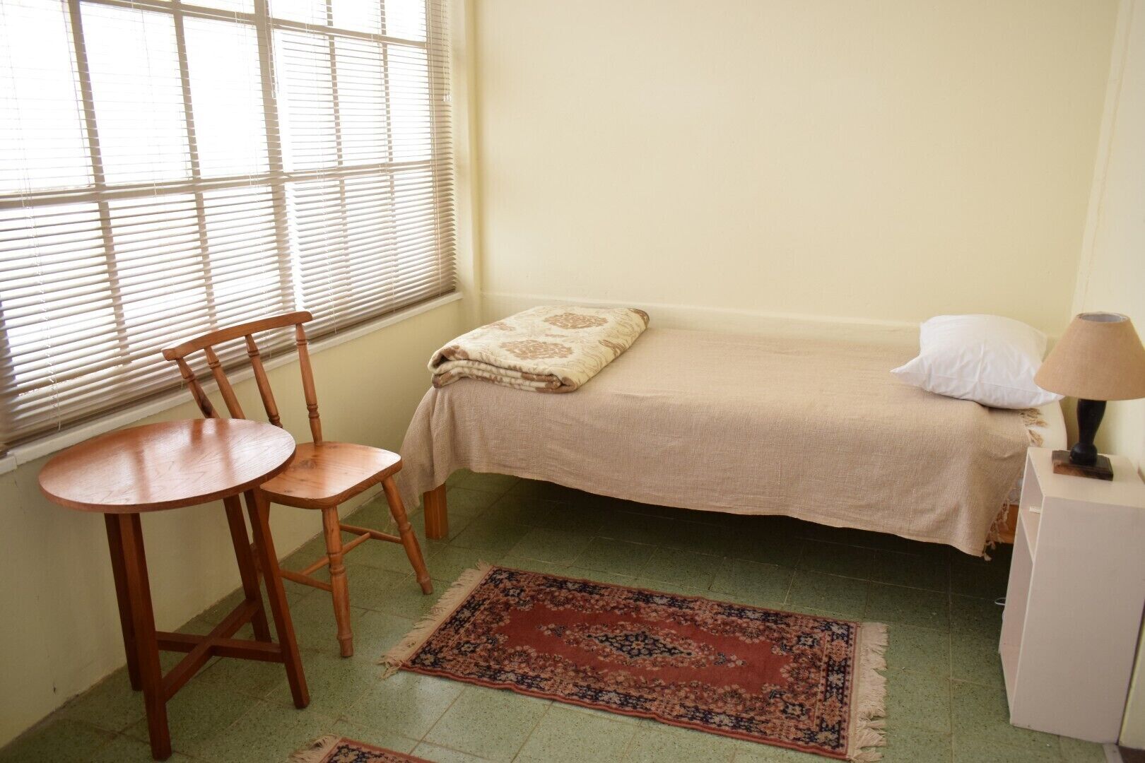 Quarto pequeno com uma cama de solteiro, uma cadeira de madeira, uma mesinha, um abajur e um tapete estampado.