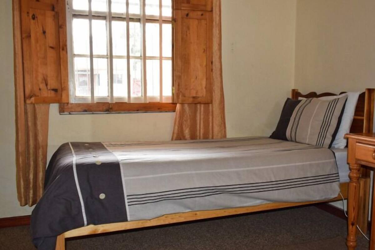 A single bed with a grey and white striped bedspread, wooden headboard, and curtains on a window.