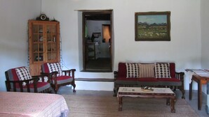 A cozy living room with a wooden ceiling, a sofa with patterned cushions, a wooden cabinet, and a painting on the wall. - Die Ou Huis (Ashton)