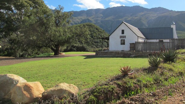 A white building with a sign reading "Die Ou Huis" in a grassy field with mountains in the background. - Die Ou Huis (Ashton)