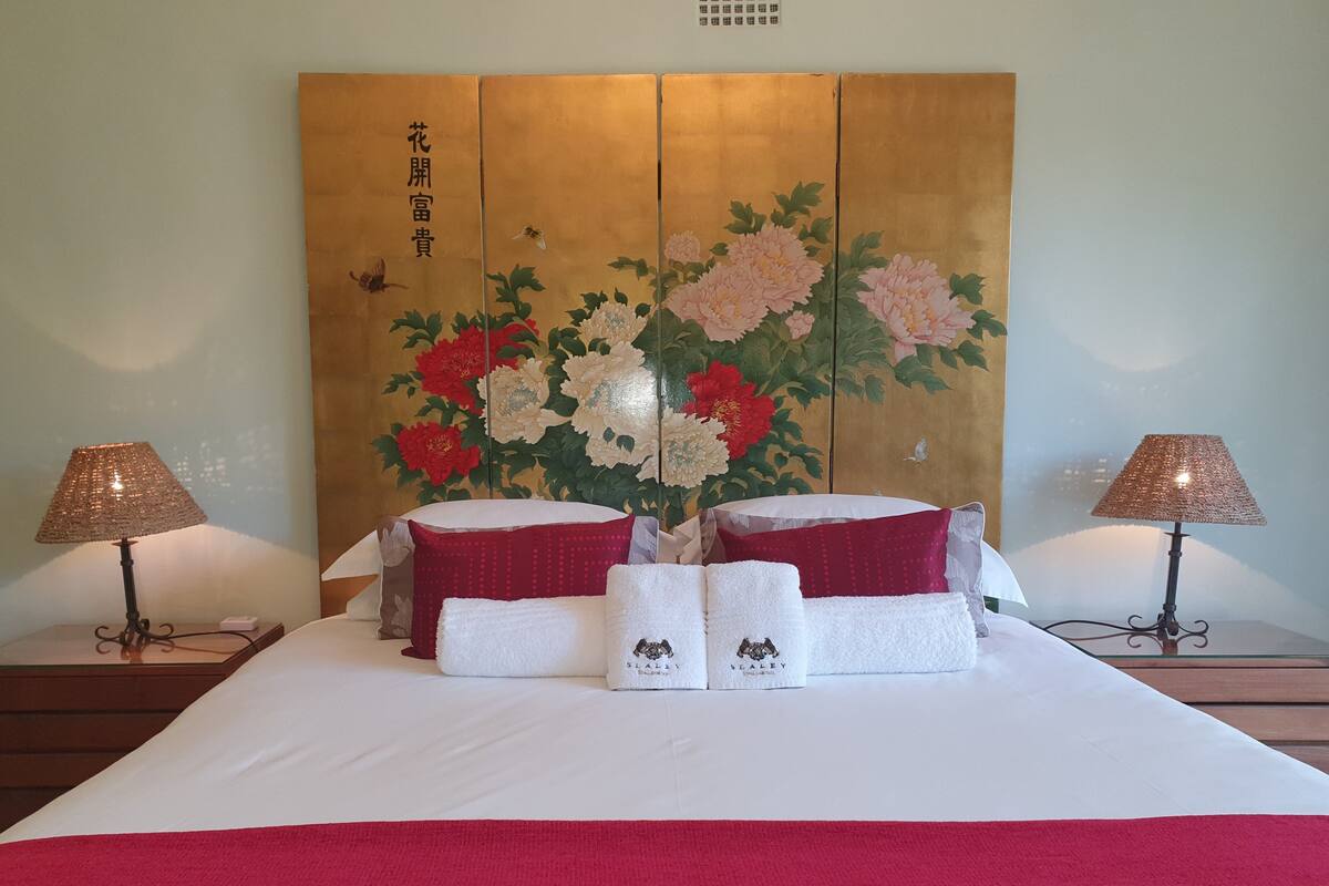 A hotel room with a large bed, two bedside lamps, a decorative headboard with floral artwork, and two towels with a logo.