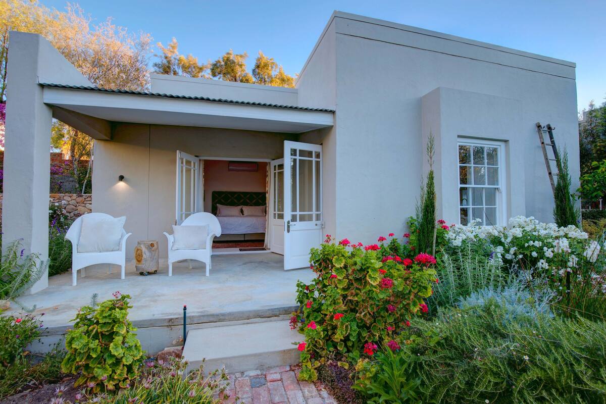 A modern house with a covered patio, white furniture, and a garden with various plants and flowers.