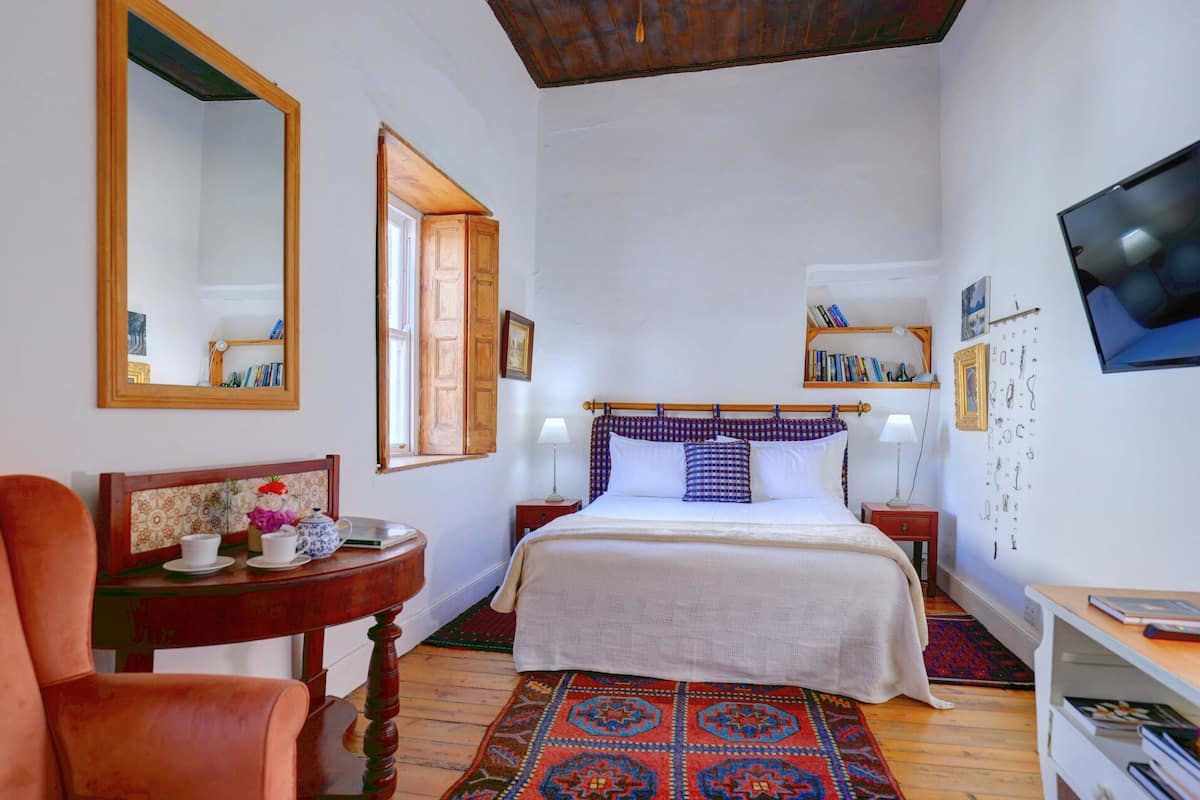 A bedroom with a large bed, a wooden chair, a small table with a teapot, a mirror, and a TV on the wall.