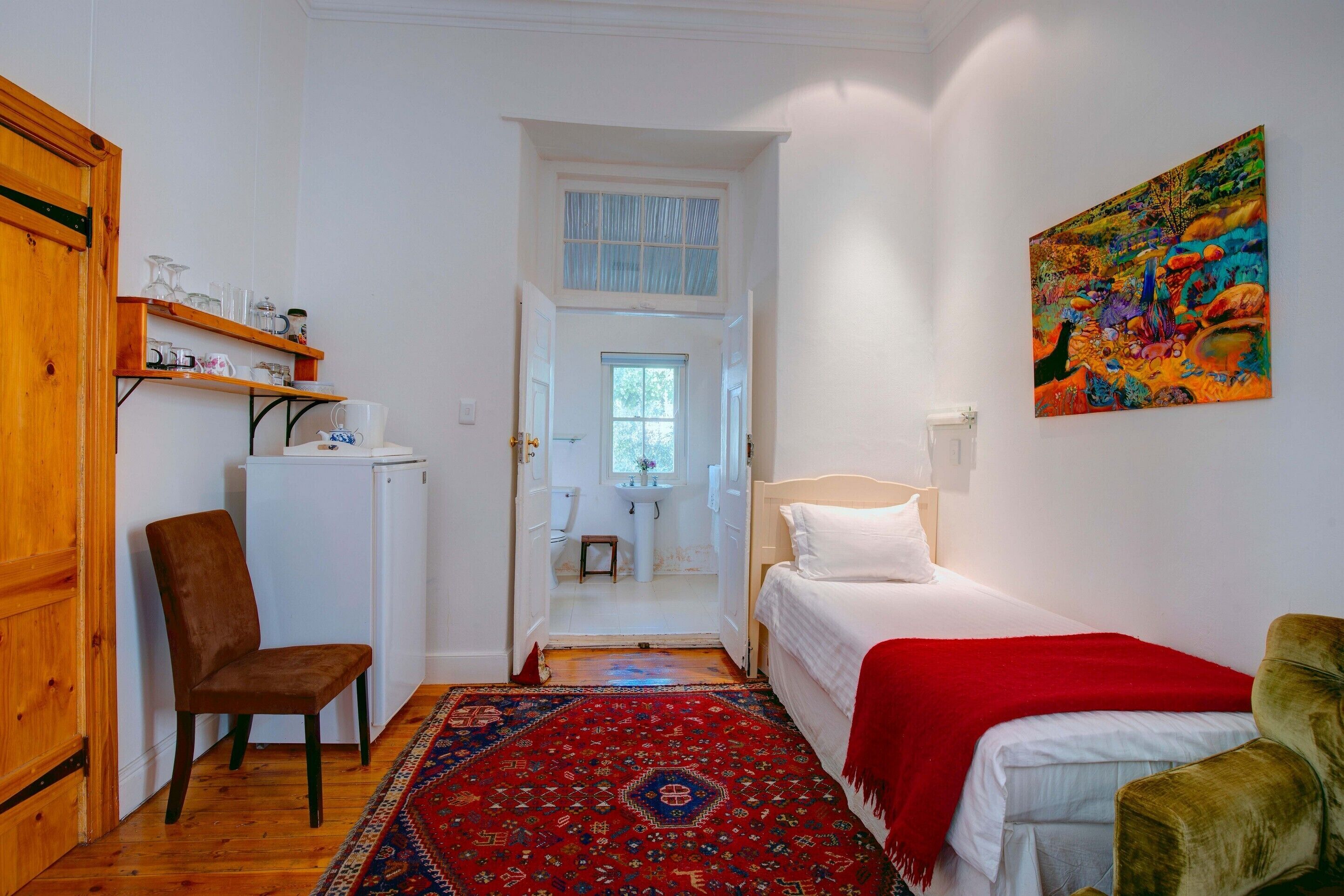 Une pièce comprenant un lit, une chaise, un bureau avec un ordinateur, un tapis rouge et un tableau accroché au mur.