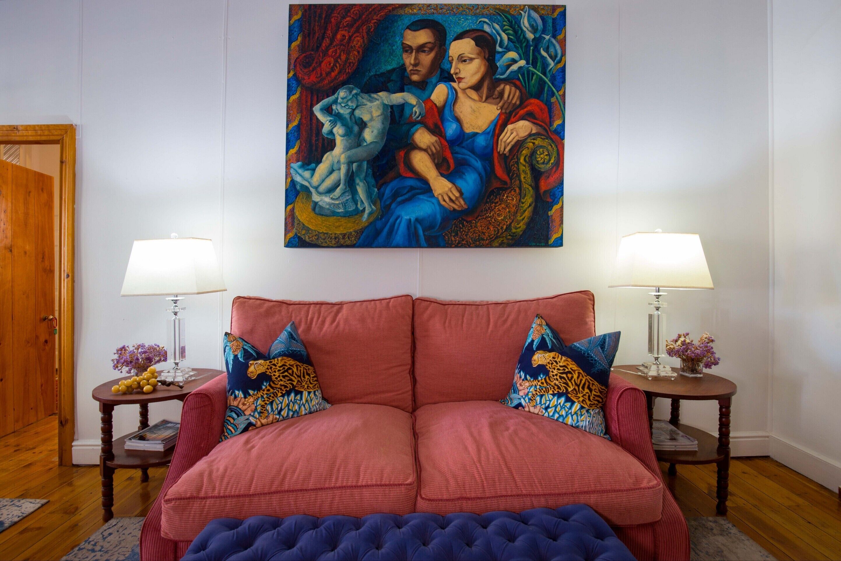 Una habitación con un sofá rojo, dos mesitas de noche con lámparas y un cuadro colorido sobre el sofá.